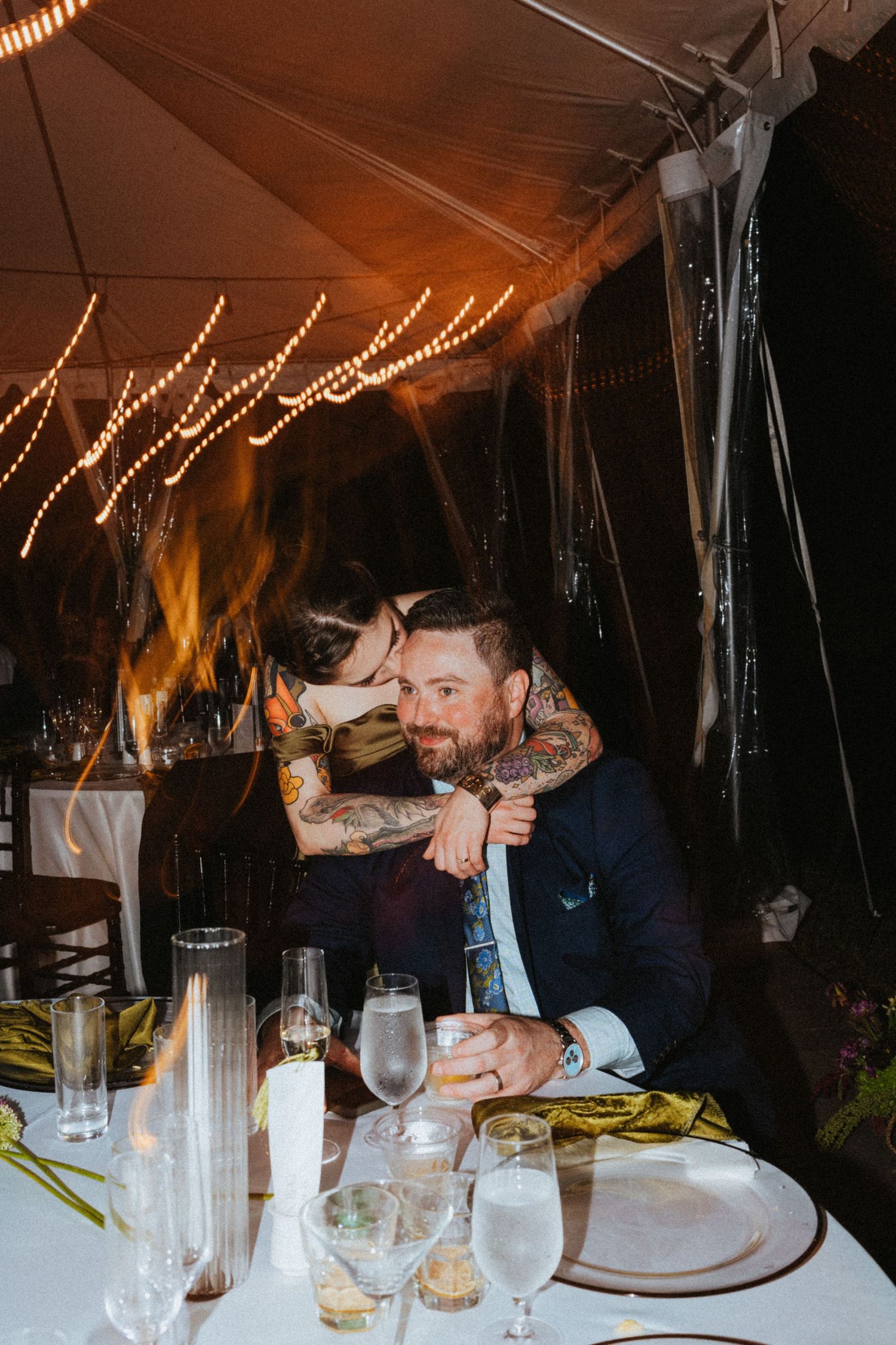 Couple embracing at wedding reception dinner table under a tent with string lights.