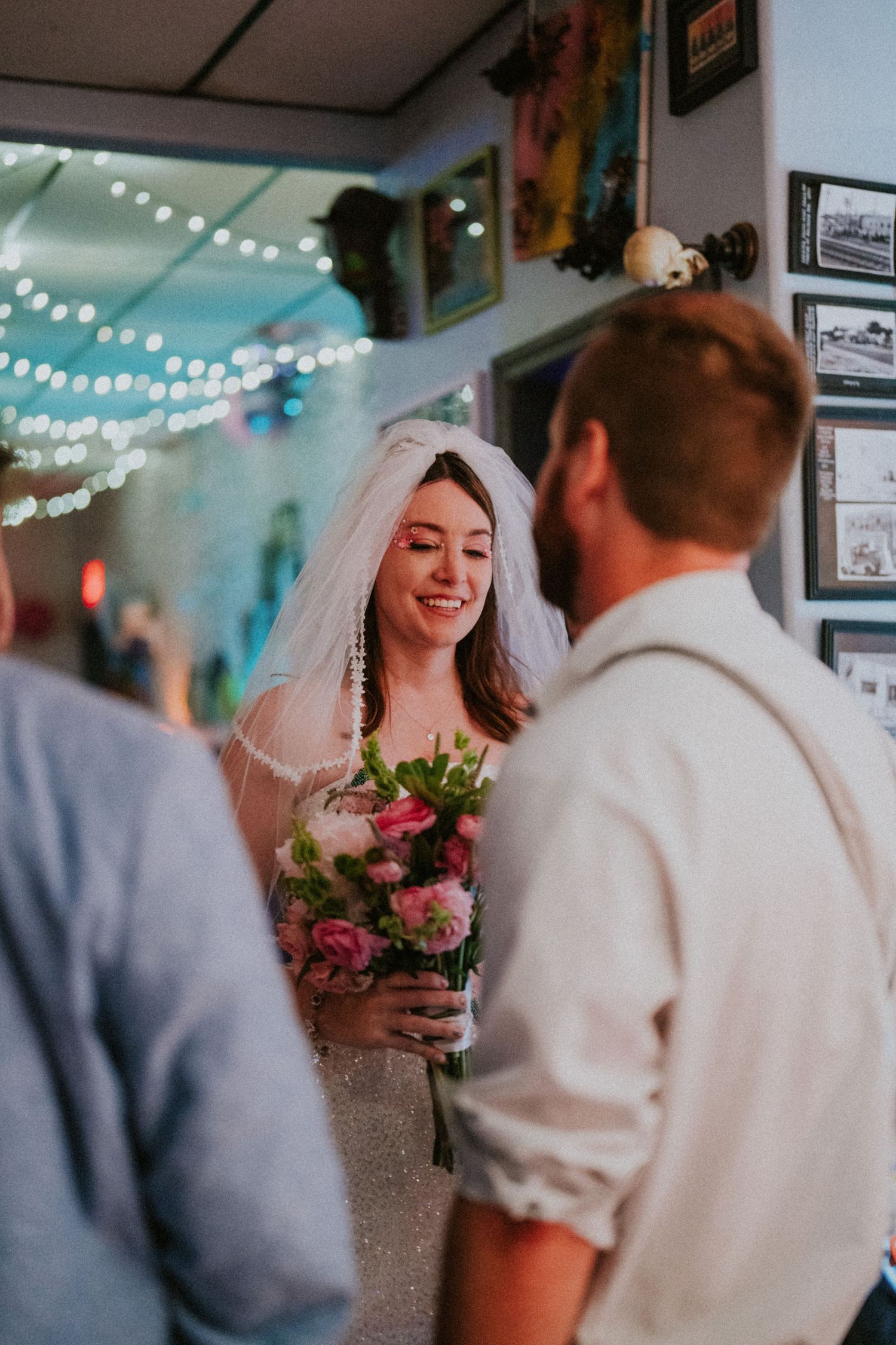 A radiant bride in a white veil holds a pink floral bouquet while smiling at guests during her wedding reception.