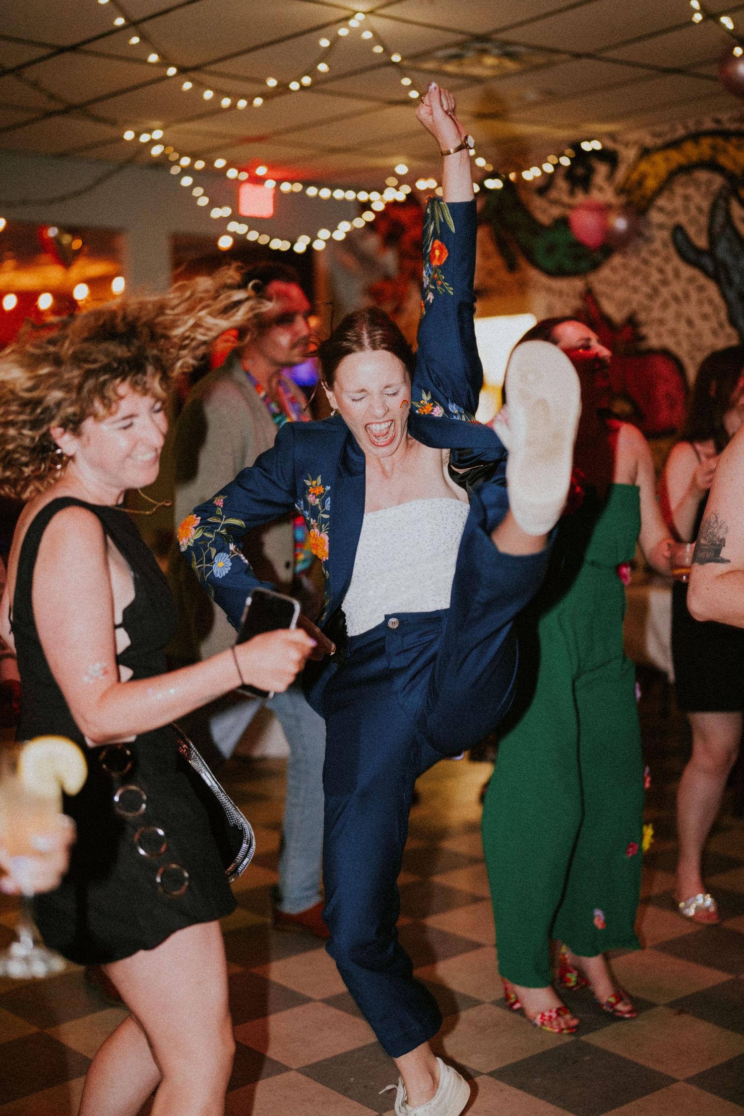 Woman in navy floral blazer dancing energetically at a party, raising her leg and arm with excitement.