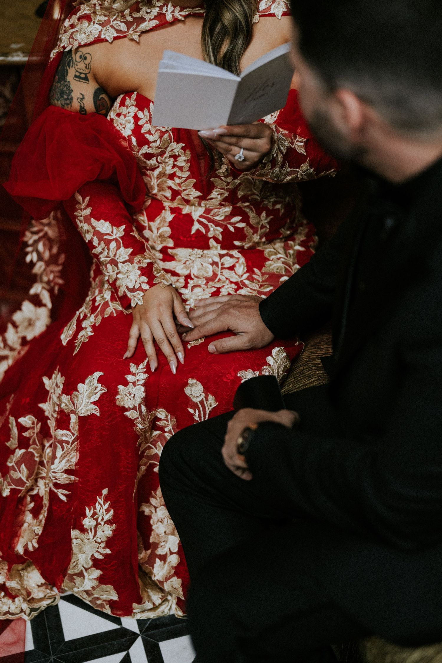 Bride in red floral gown holding hands with groom in black suit while reading wedding vows.