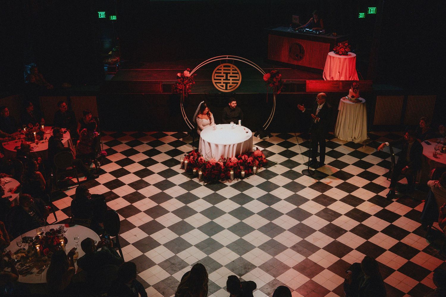 Couple at elegant wedding reception on checkered dance floor with red roses and circular backdrop.