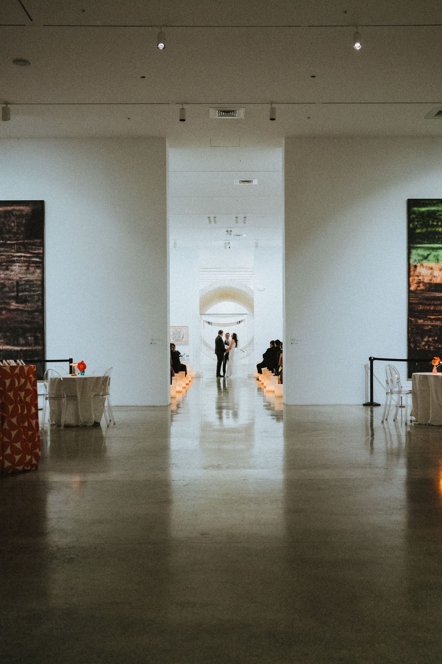 Elegant wedding ceremony in a modern art museum gallery with candlelit aisle and couple silhouetted in arched doorway.