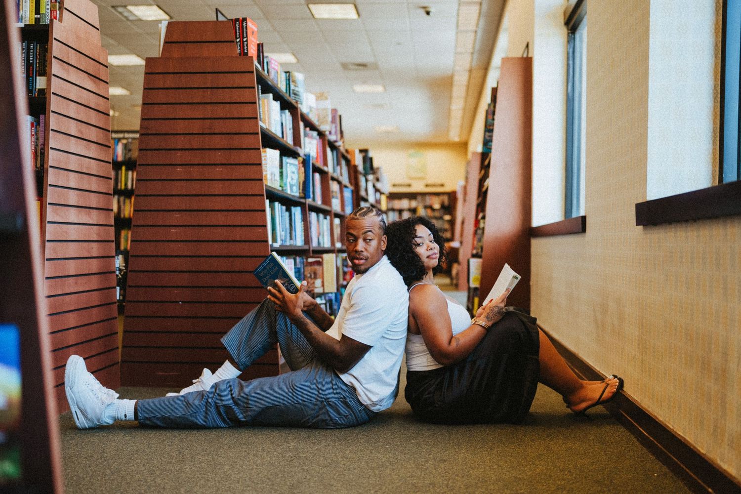 Couple sitting on bookstore floor reading books back-to-back between colorful bookshelves.