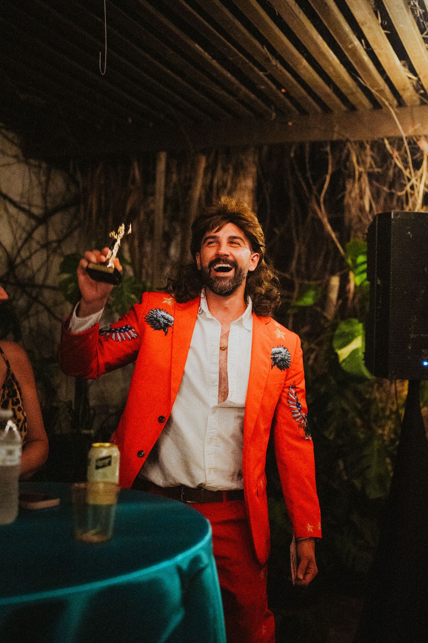 Man laughing in bold orange embroidered suit holding a trophy at a festive indoor celebration event.