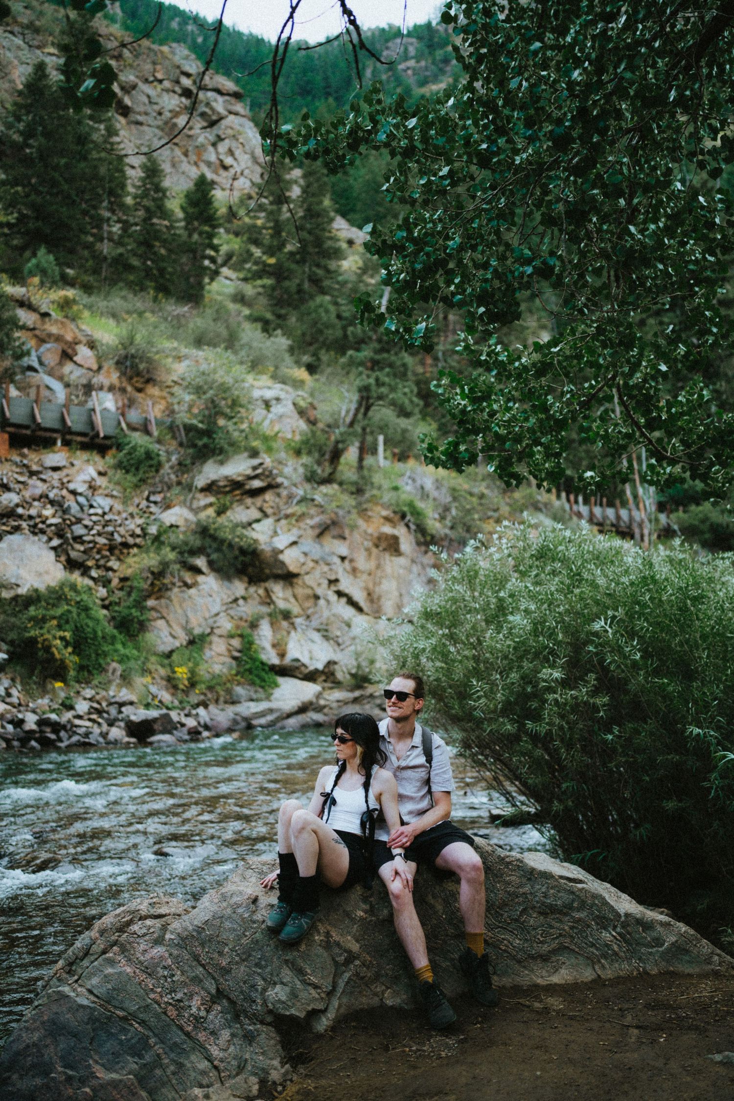 Couple sitting on a boulder beside a rushing mountain river surrounded by lush green trees.