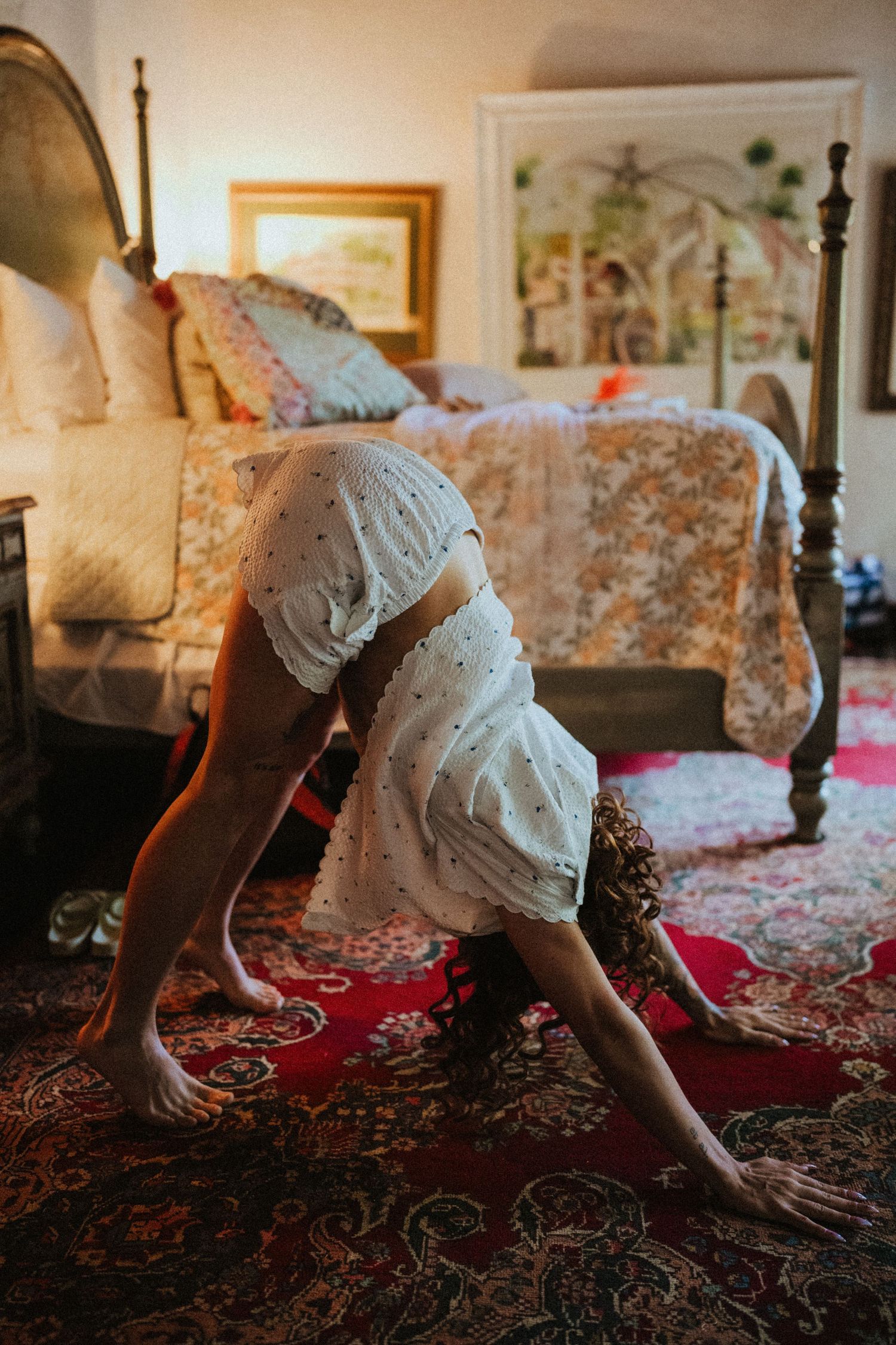 Woman in white dress doing a forward fold yoga pose on a red patterned rug in a cozy bedroom.