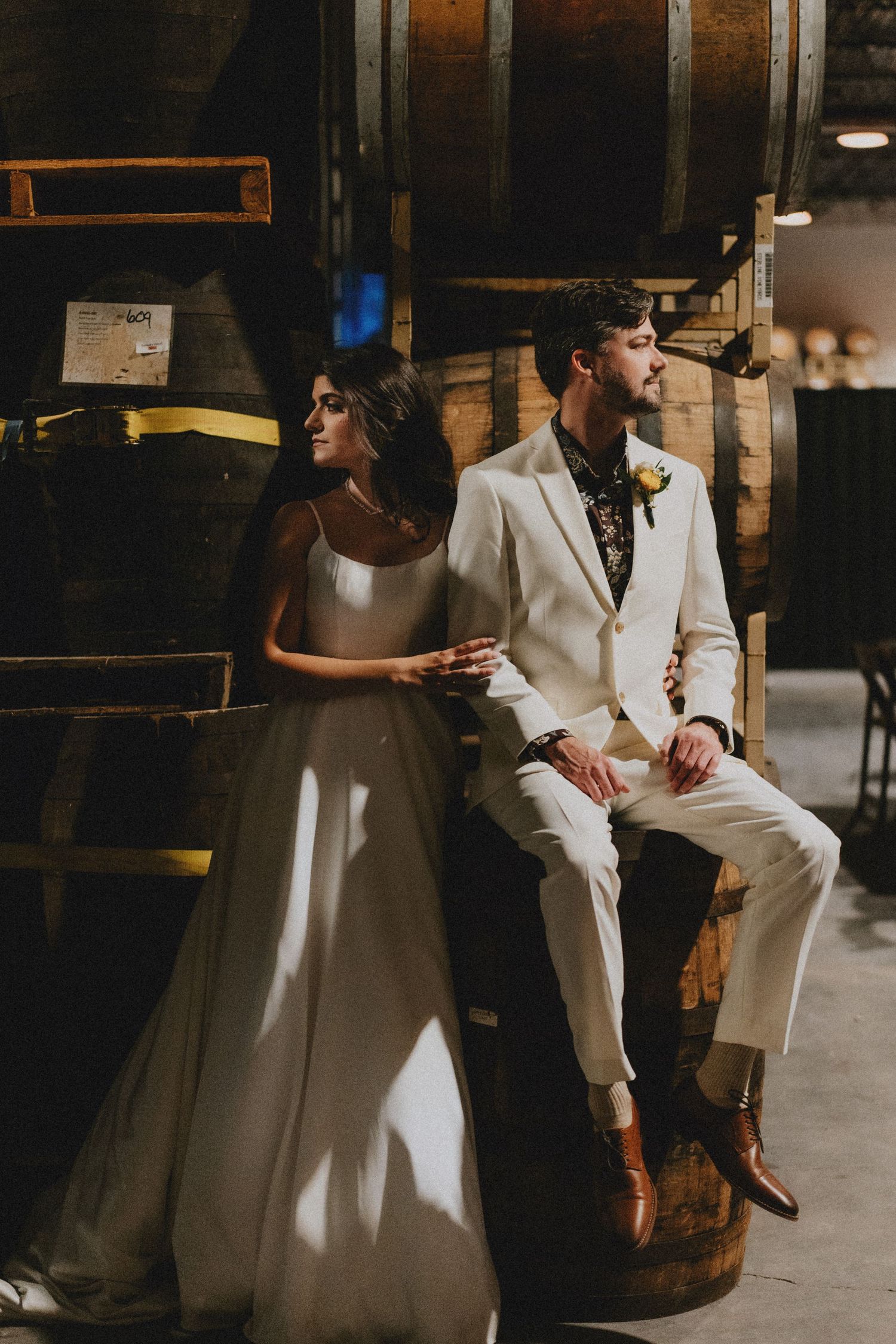 Bride in white gown and groom in ivory suit pose among oak barrels in a rustic winery wedding venue.