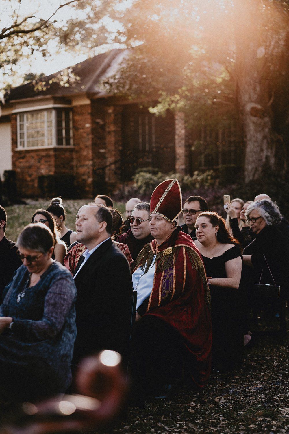 Wedding guests gathered outdoors at sunset with brick building in background.
