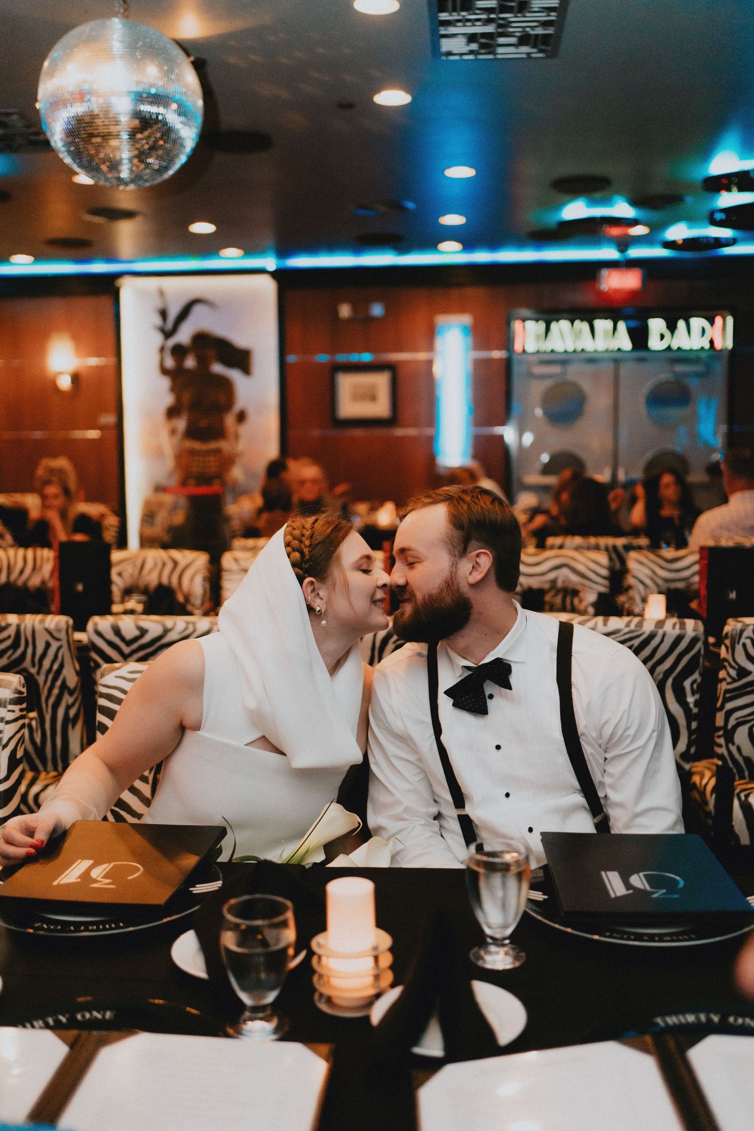 Bride and groom share a romantic kiss at their wedding reception in a retro bar with zebra print booths and disco ball.