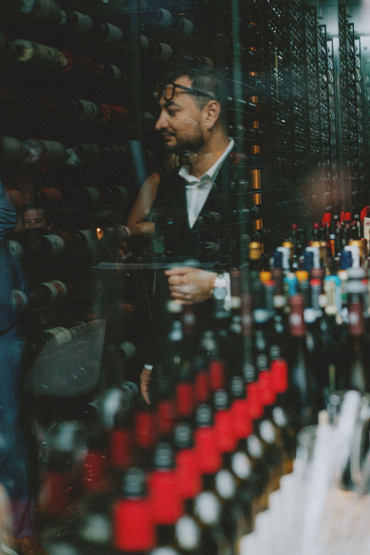 Man browsing extensive wine cellar collection viewed through glass wall reflection.