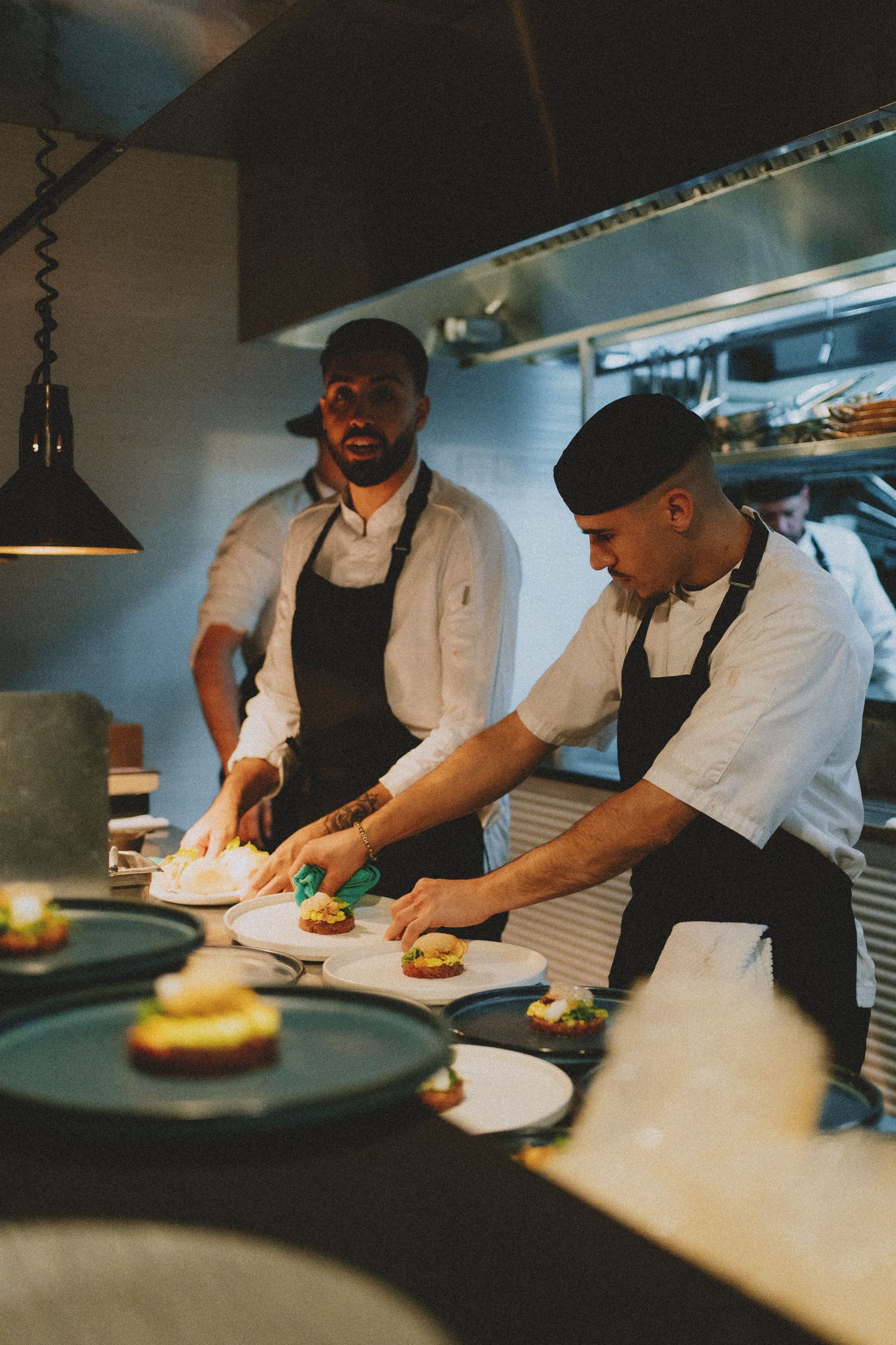 Two chefs in black aprons carefully plate gourmet dishes in a professional restaurant kitchen.