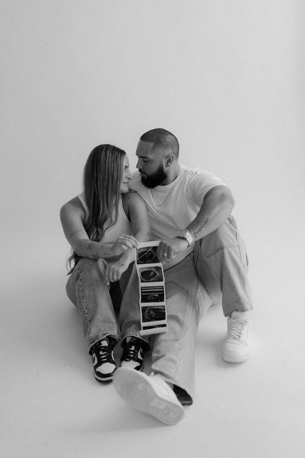 A couple sits intimately on a white backdrop holding ultrasound photos, suggesting a pregnancy announcement.