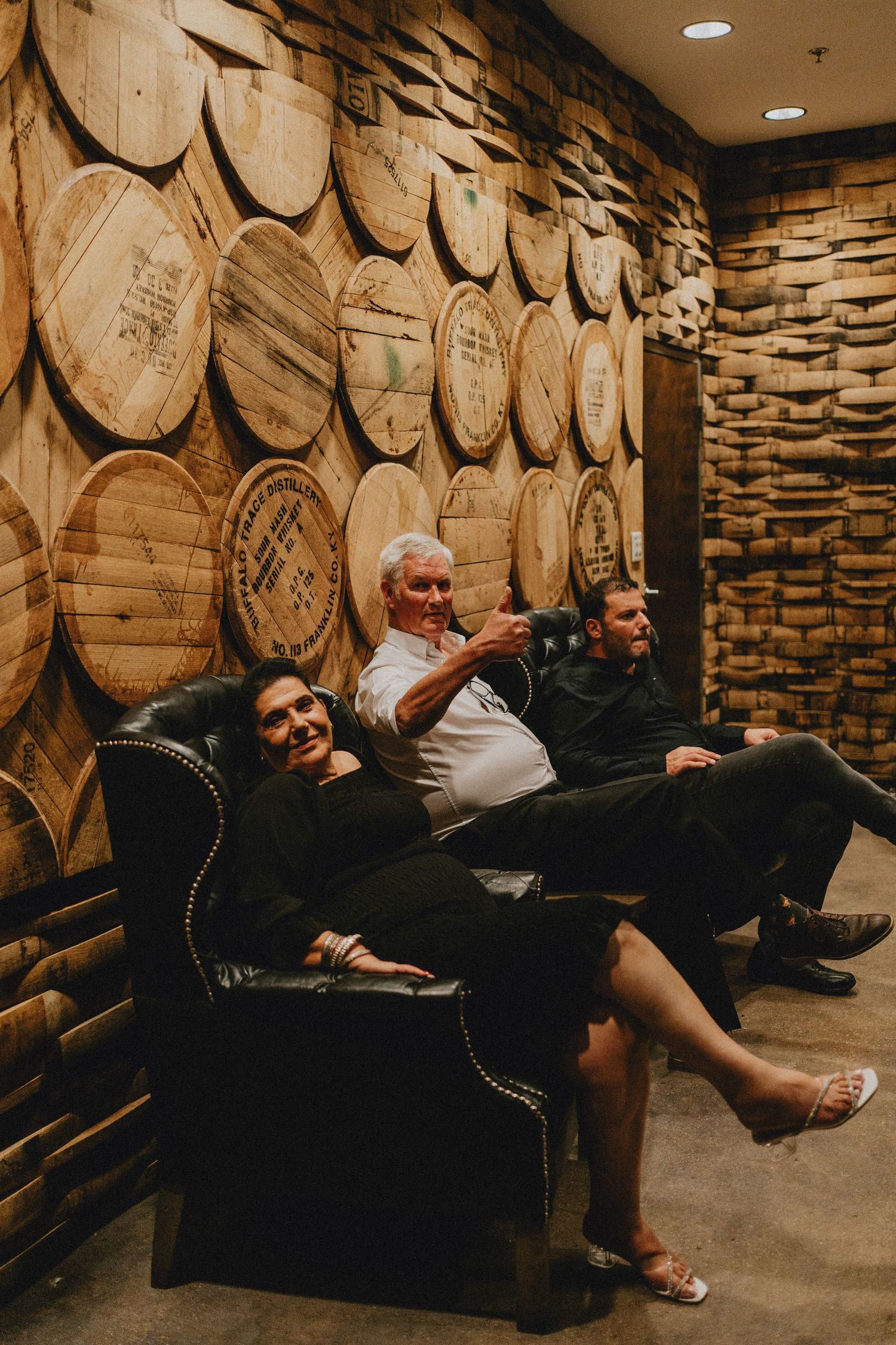 Three people relaxing on a leather chair in front of a wall of wooden barrel lids.