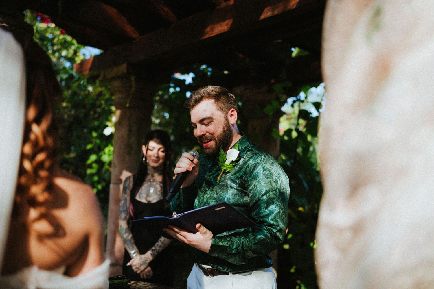A tattooed officiant reads vows at an outdoor wedding ceremony surrounded by lush greenery.