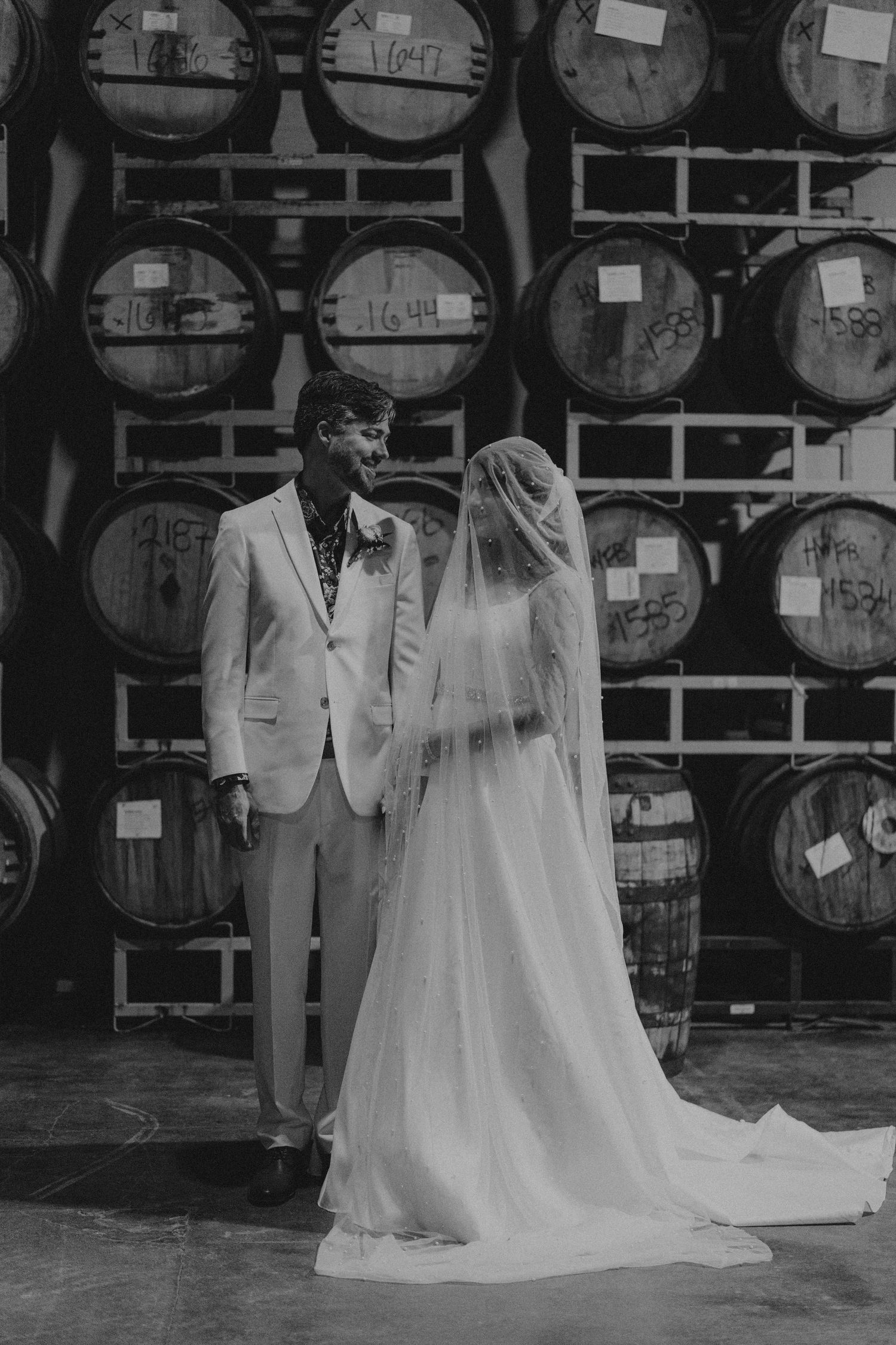 Black and white photo of bride and groom sharing a moment in front of wine barrels at a winery wedding venue.