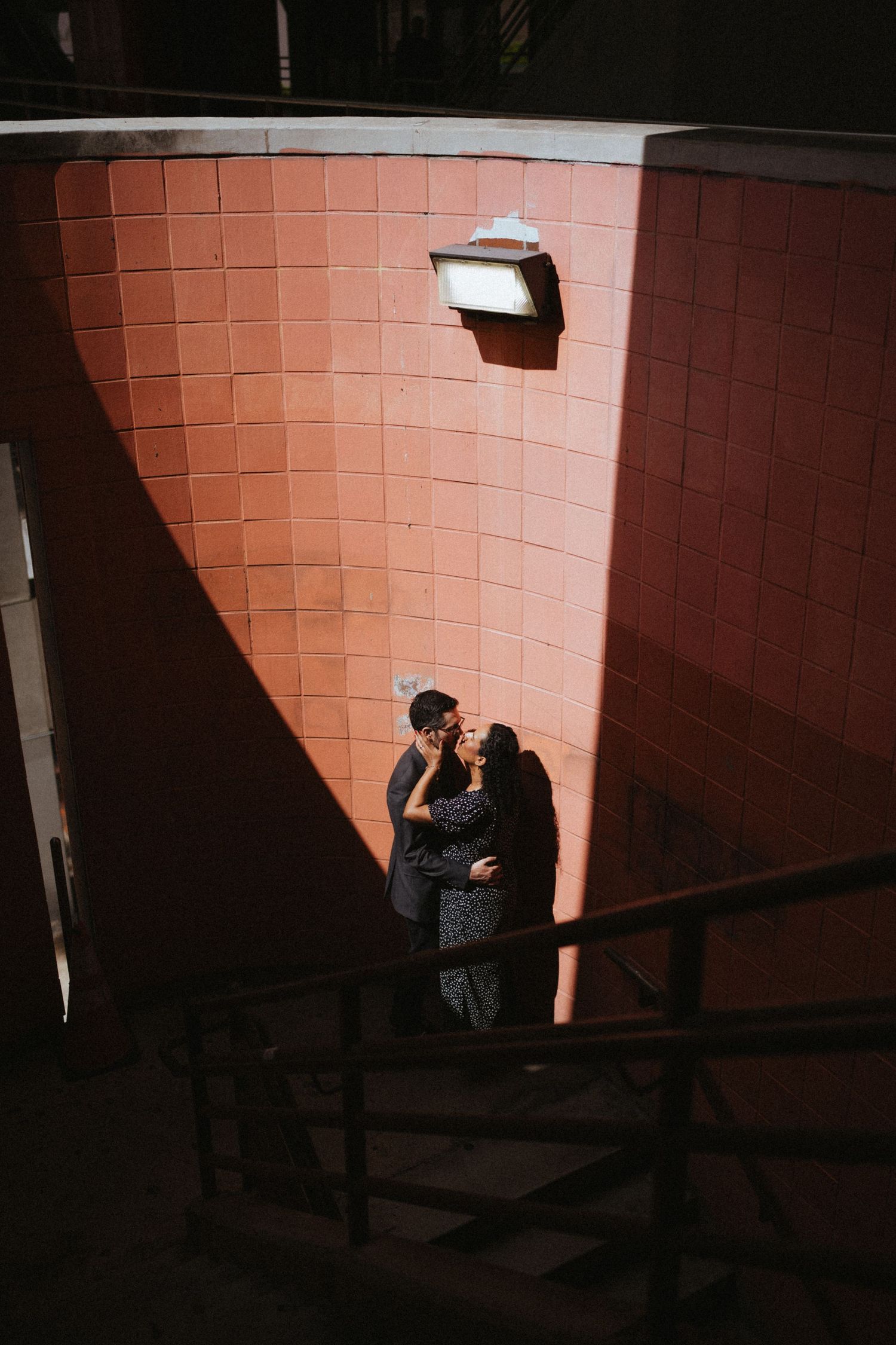 Couple embracing romantically in sunlit corner of red brick wall near outdoor staircase.