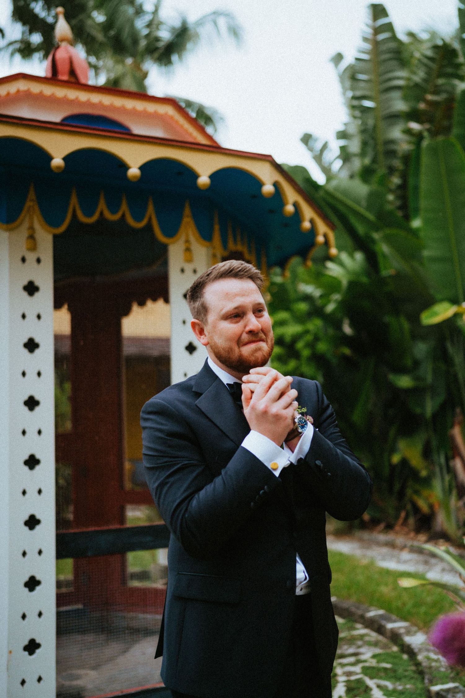 Groom in black suit adjusting cufflinks near colorful gazebo surrounded by tropical plants.