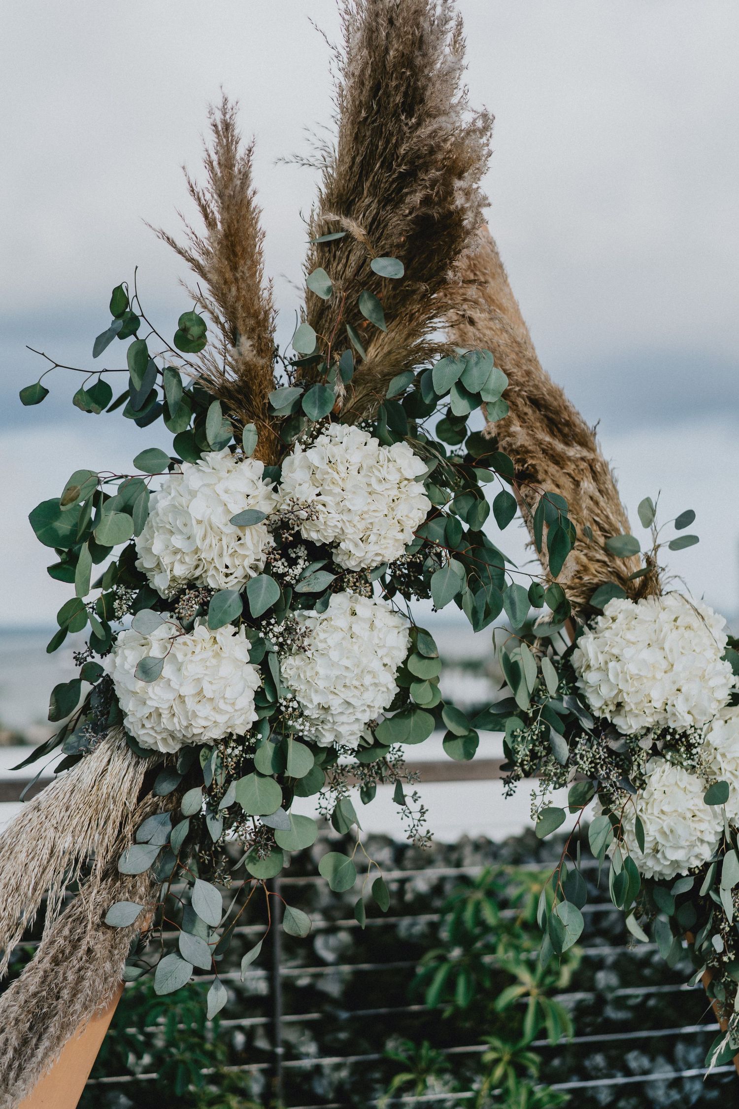 Boho wedding arch with white hydrangeas, eucalyptus, and pampas grass against an overcast sky.