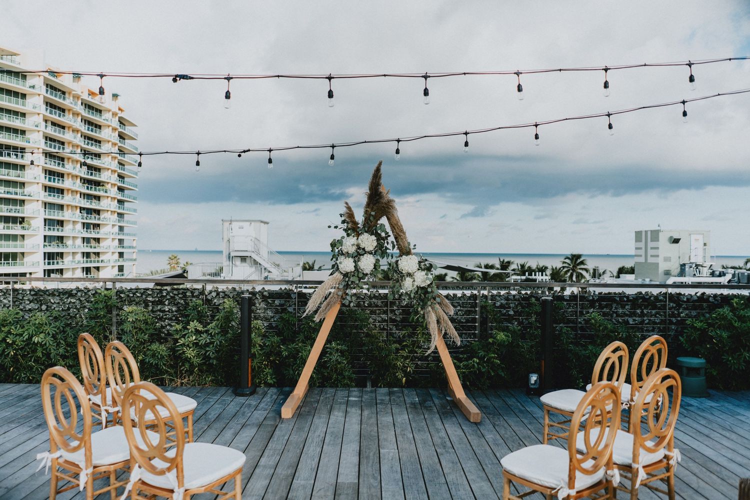 Rooftop wedding ceremony setup with wooden arch, floral decor, rattan chairs, string lights, and city skyline backdrop.