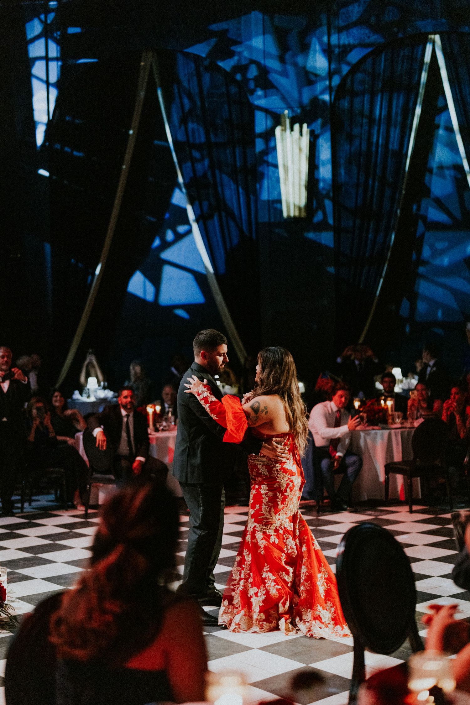 Couple sharing first dance on checkered floor in elegant venue, bride in red floral gown, guests seated at candlelit tables.