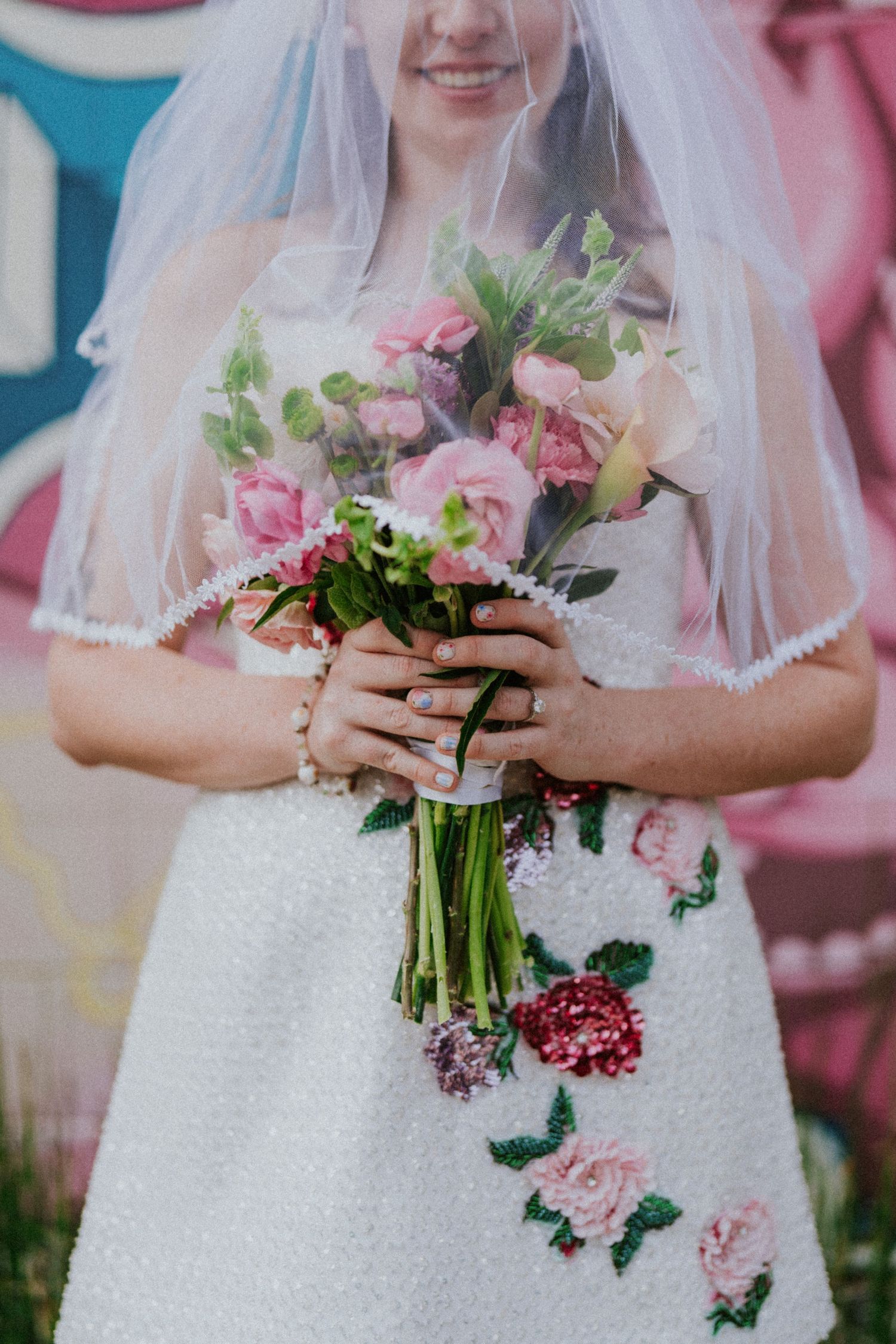 Close-up of a bride in a white floral embroidered mini dress holding a pink and green wedding bouquet with a lace veil.