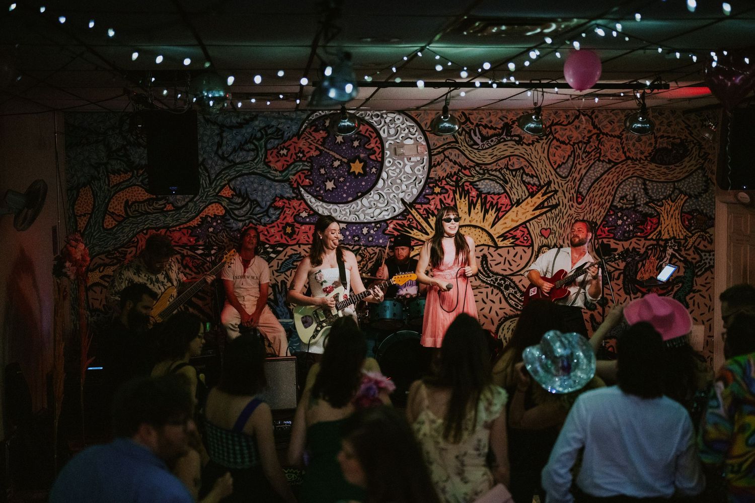 Live band performing on stage with colorful psychedelic mural backdrop, string lights, and dancing crowd in a cozy venue.