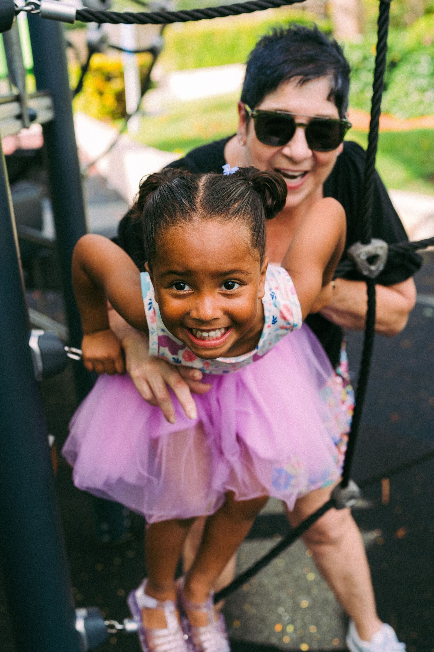 A smiling young girl in a pink tutu plays on playground equipment while a woman in sunglasses assists her from behind.