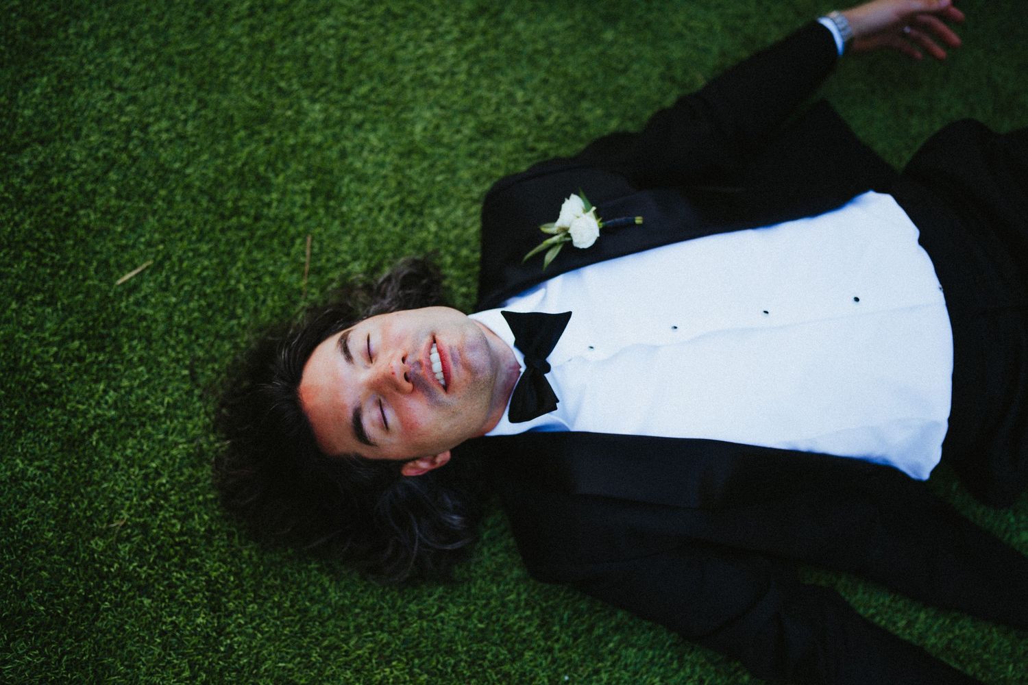 Man in black tuxedo with white boutonniere lying on green grass, smiling with eyes closed.