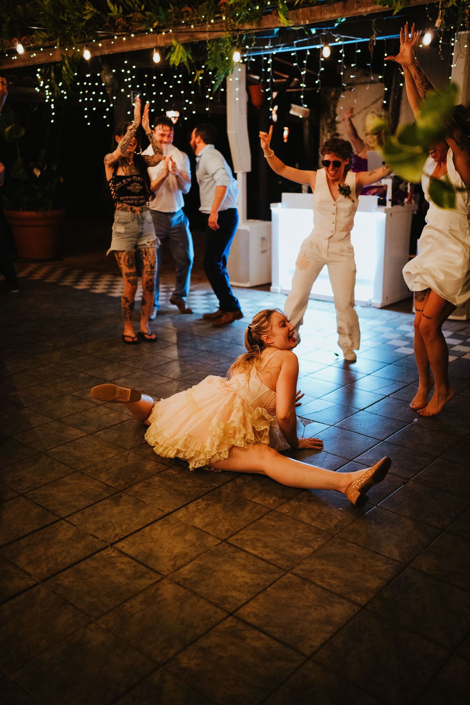 Young girl in white dress does the splits on a dance floor at a festive outdoor wedding reception with string lights.