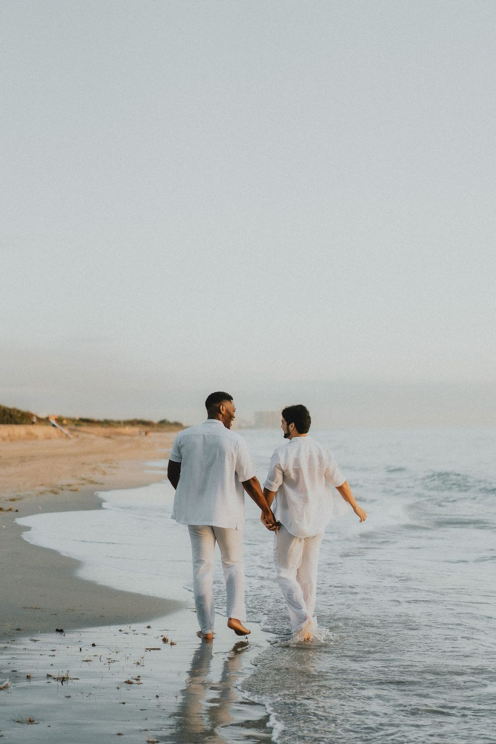 A couple in white outfits walking together along a misty beach shoreline.