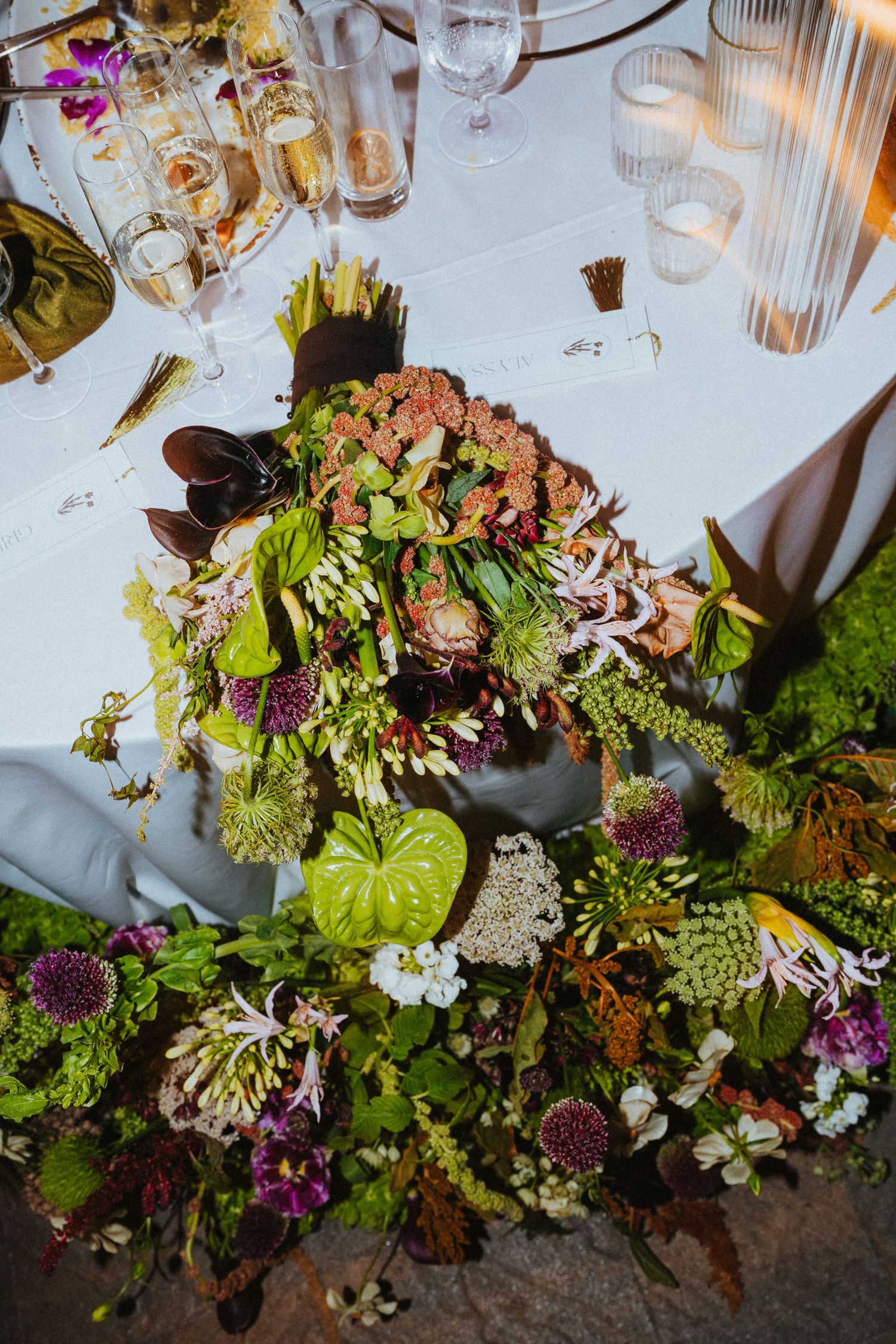 Lush wedding table centerpiece featuring green anthuriums, burgundy blooms, and wildflowers on a white linen tablecloth.
