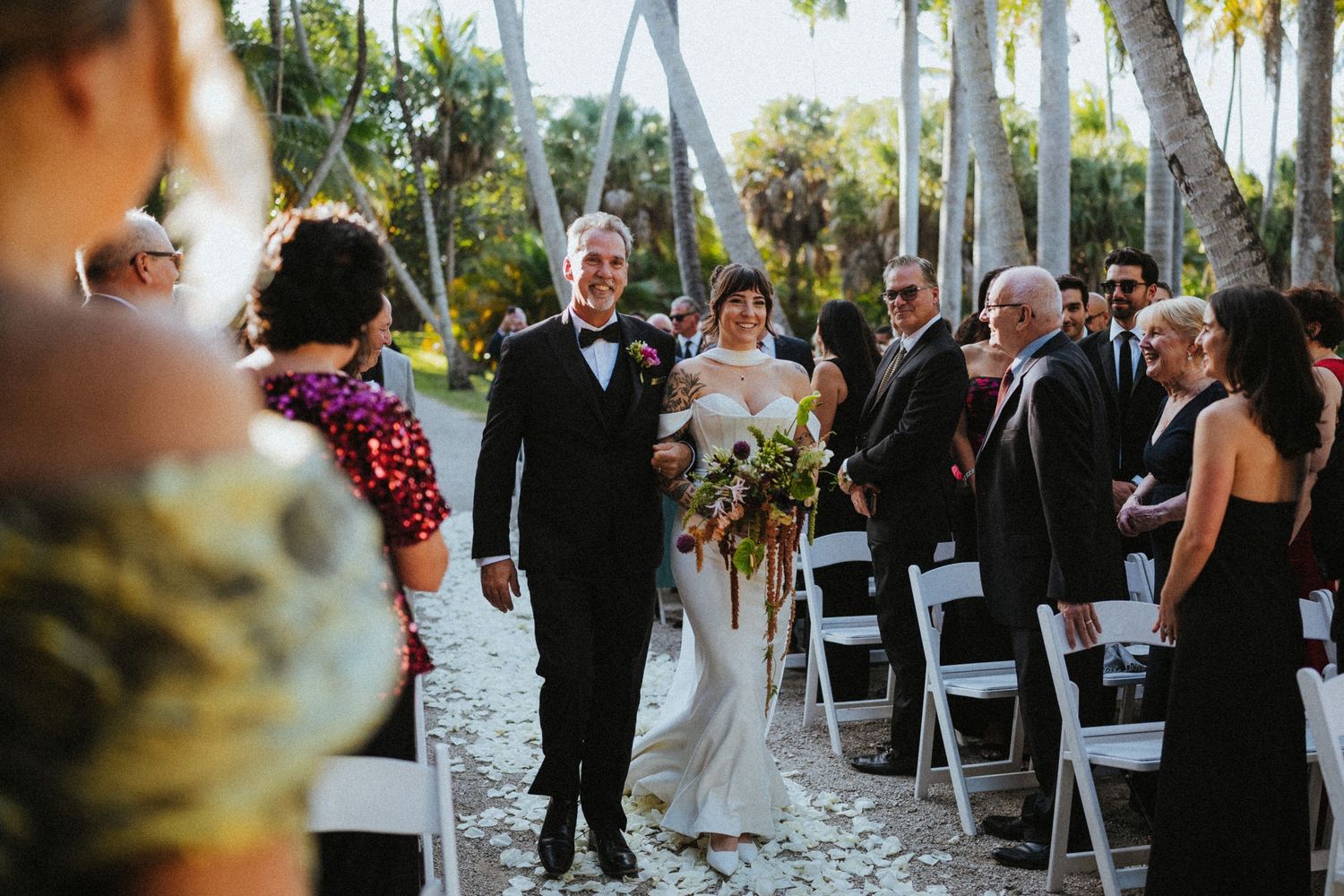 Bride and groom walk down outdoor aisle lined with white chairs and palm trees at a tropical wedding ceremony.