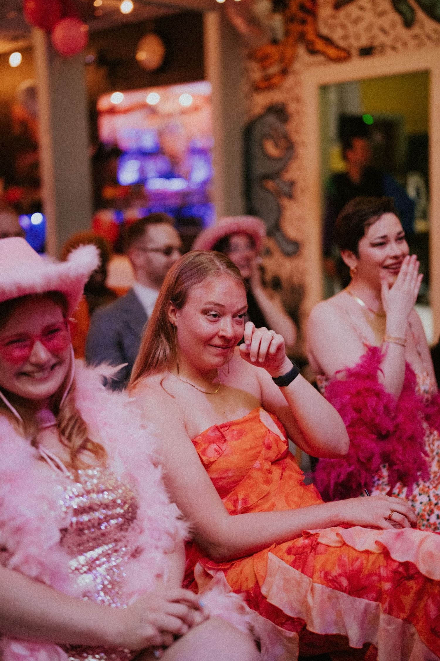 Women in pink feather boas and cowgirl hats laugh at a lively themed party with colorful decor.