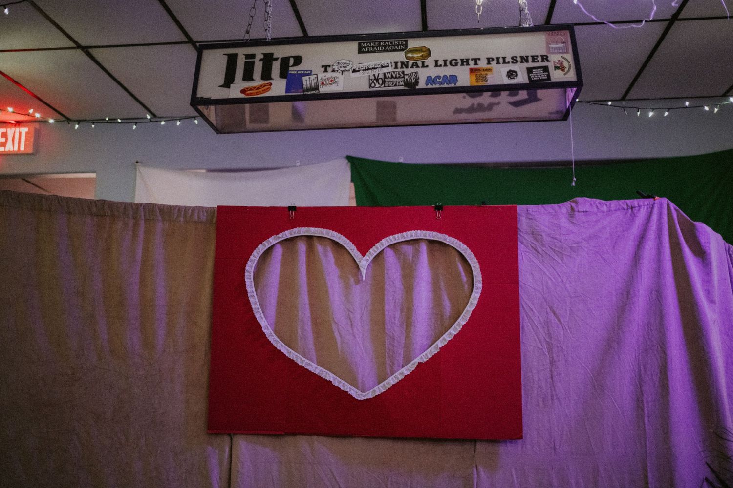 Red fabric banner with white heart outline hanging on wall beneath Miller Lite sign in bar setting.