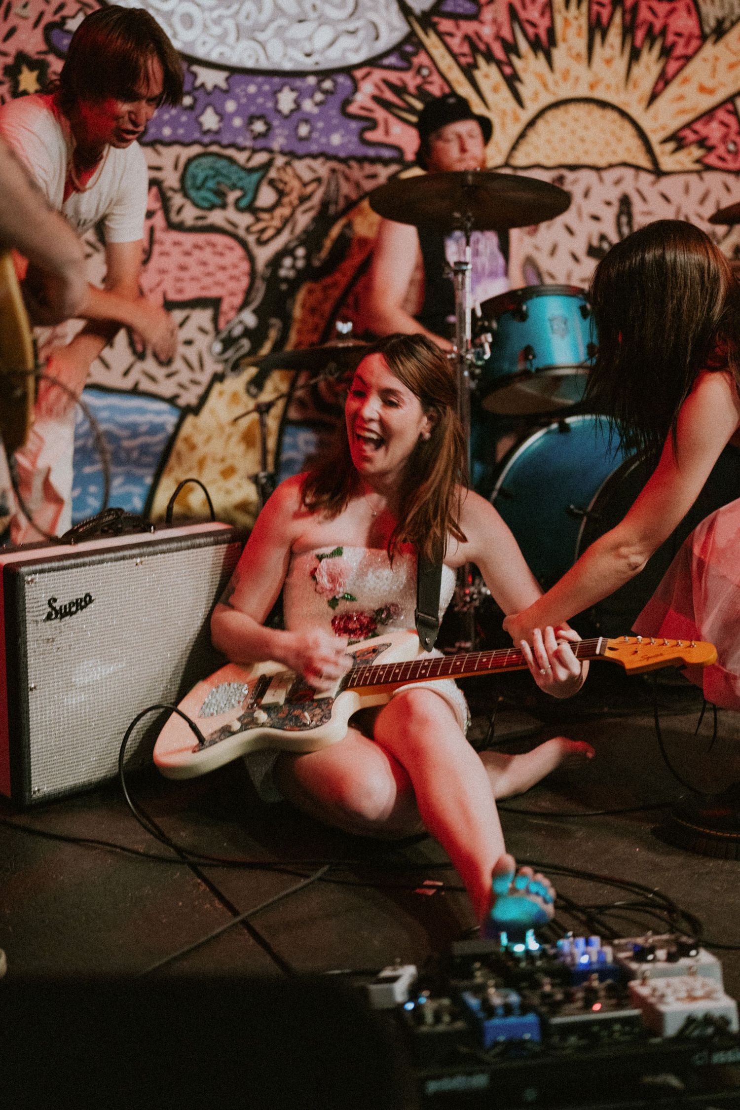 Bride in floral dress sits on stage floor laughing while playing electric guitar at wedding reception.