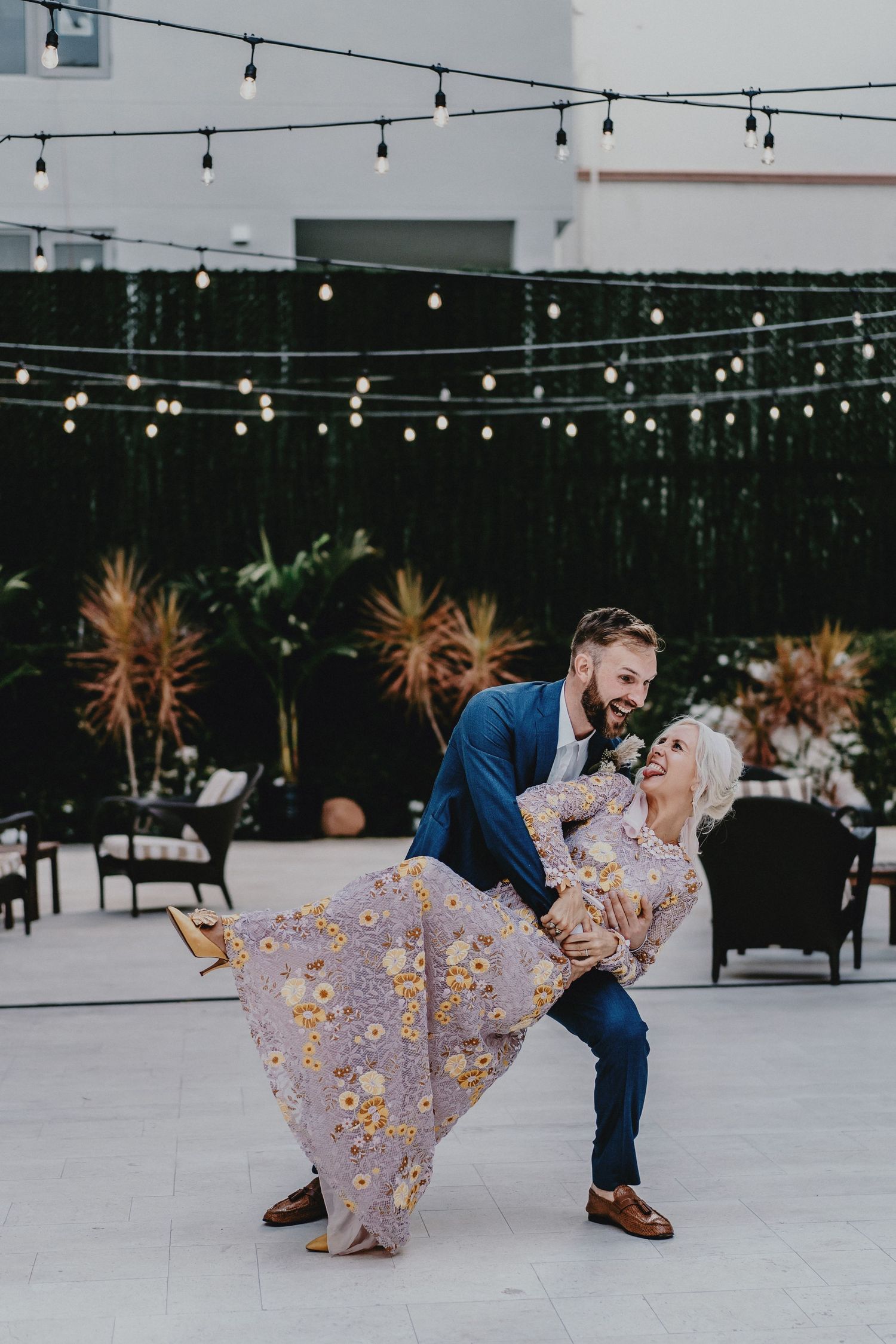 A couple dances outdoors under string lights, the woman in a floral dress being dipped by a man in a blue suit.