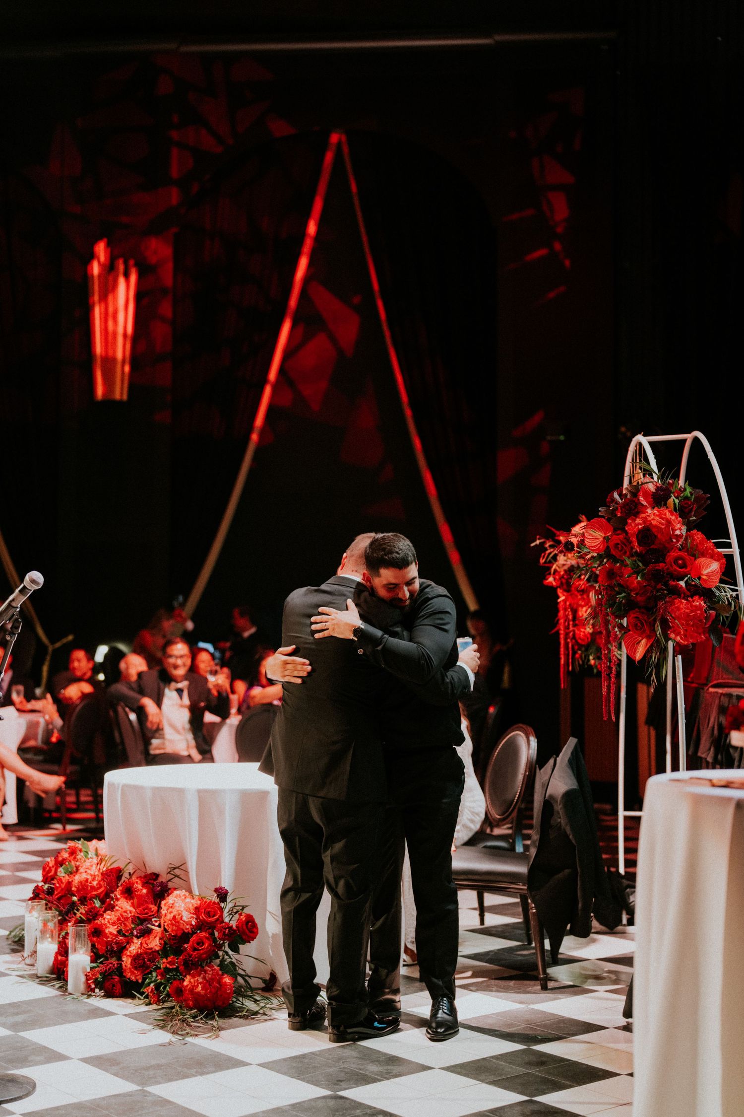 Two men in black tuxedos sharing an emotional embrace at a wedding reception with red floral centerpieces.