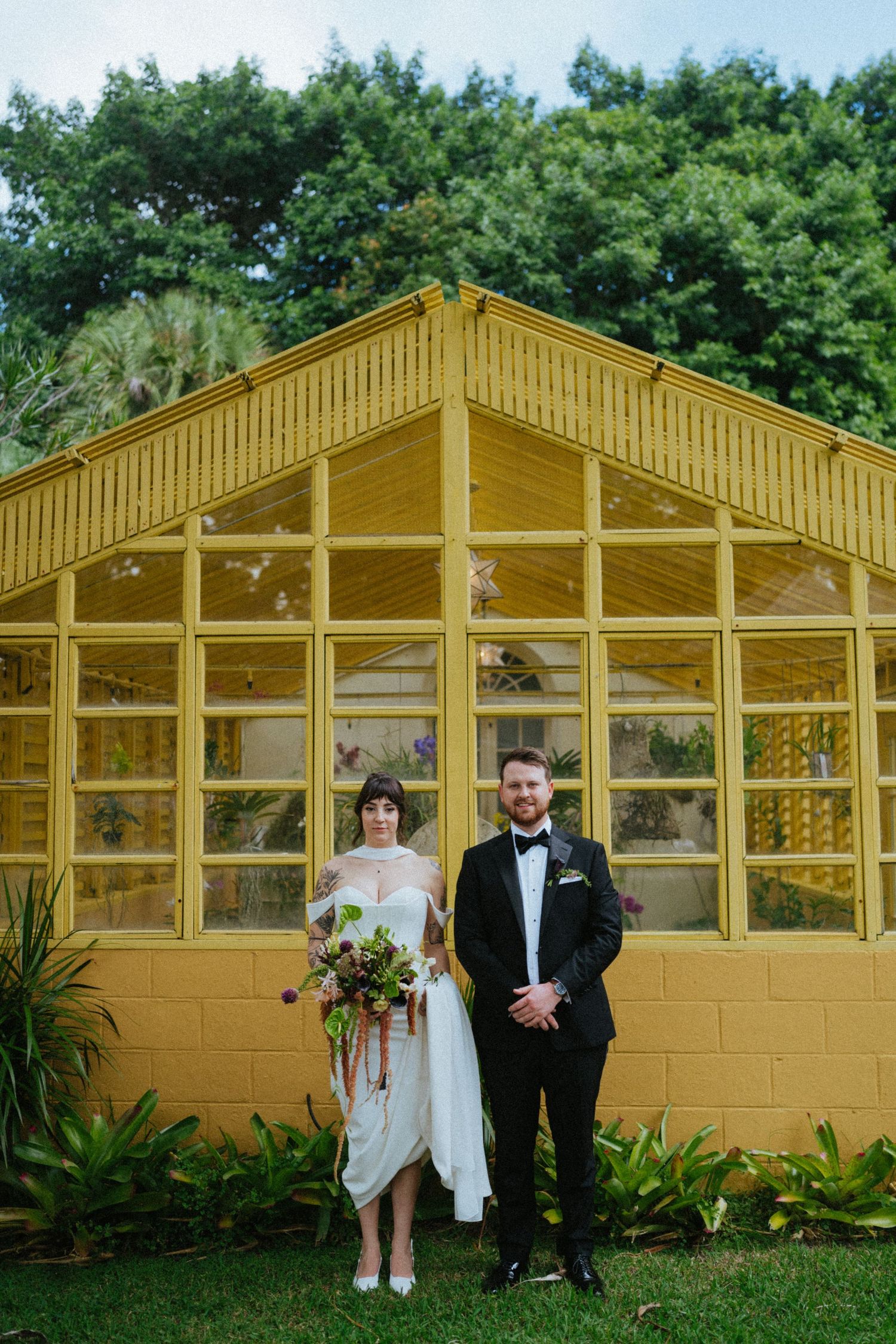 Bride and groom stand before a yellow greenhouse surrounded by lush greenery on their wedding day.