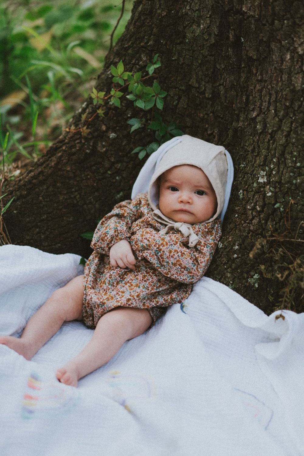 Baby wearing floral dress and white bonnet rests on blanket against tree trunk in outdoor garden setting.