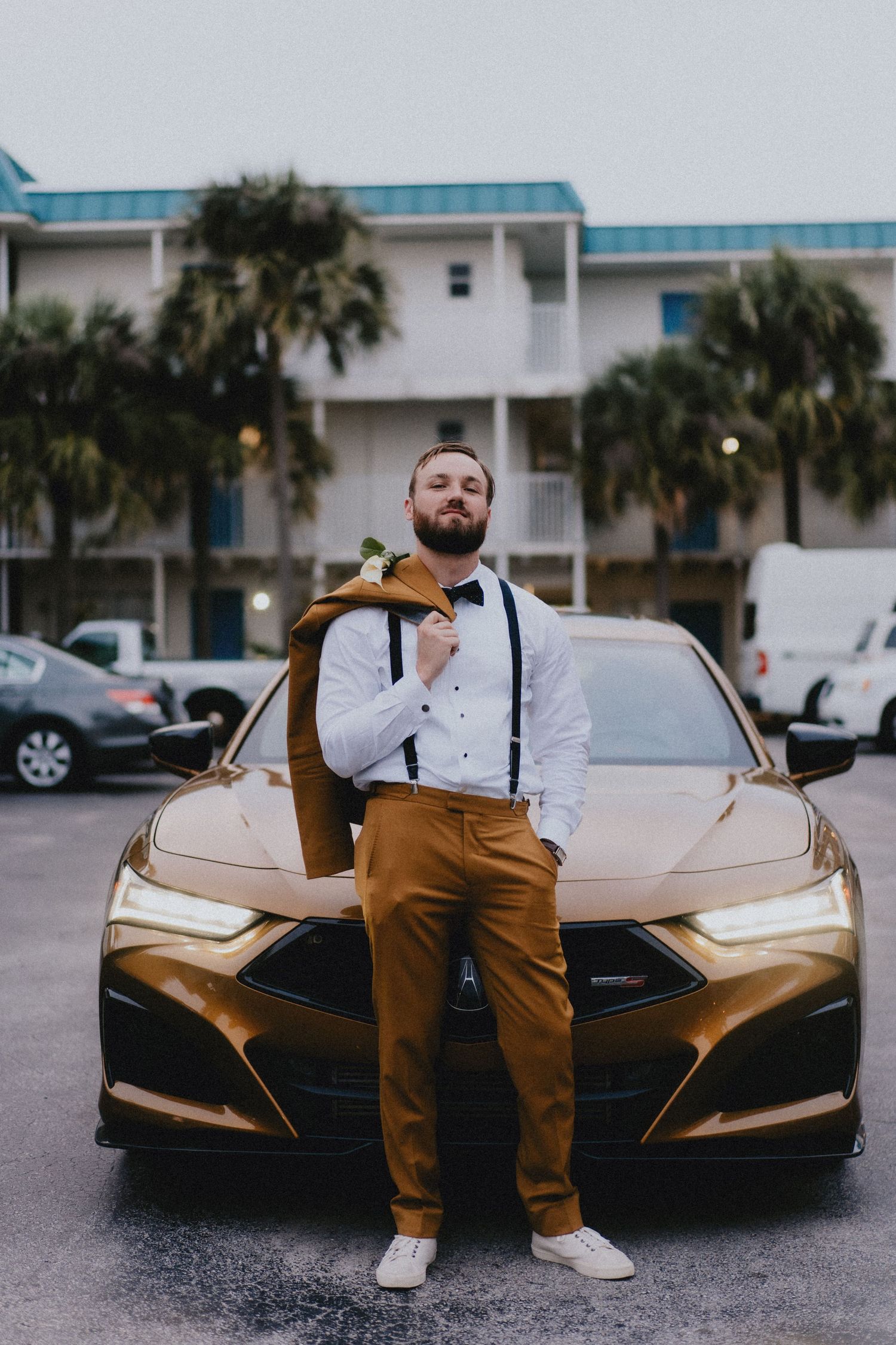 Stylish groom in mustard pants, white shirt, suspenders, and bow tie poses beside a gold car with palm trees.