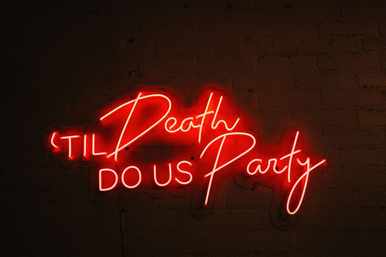Red neon sign reading 'Til Death Do Us Party' mounted on a dark brick wall.