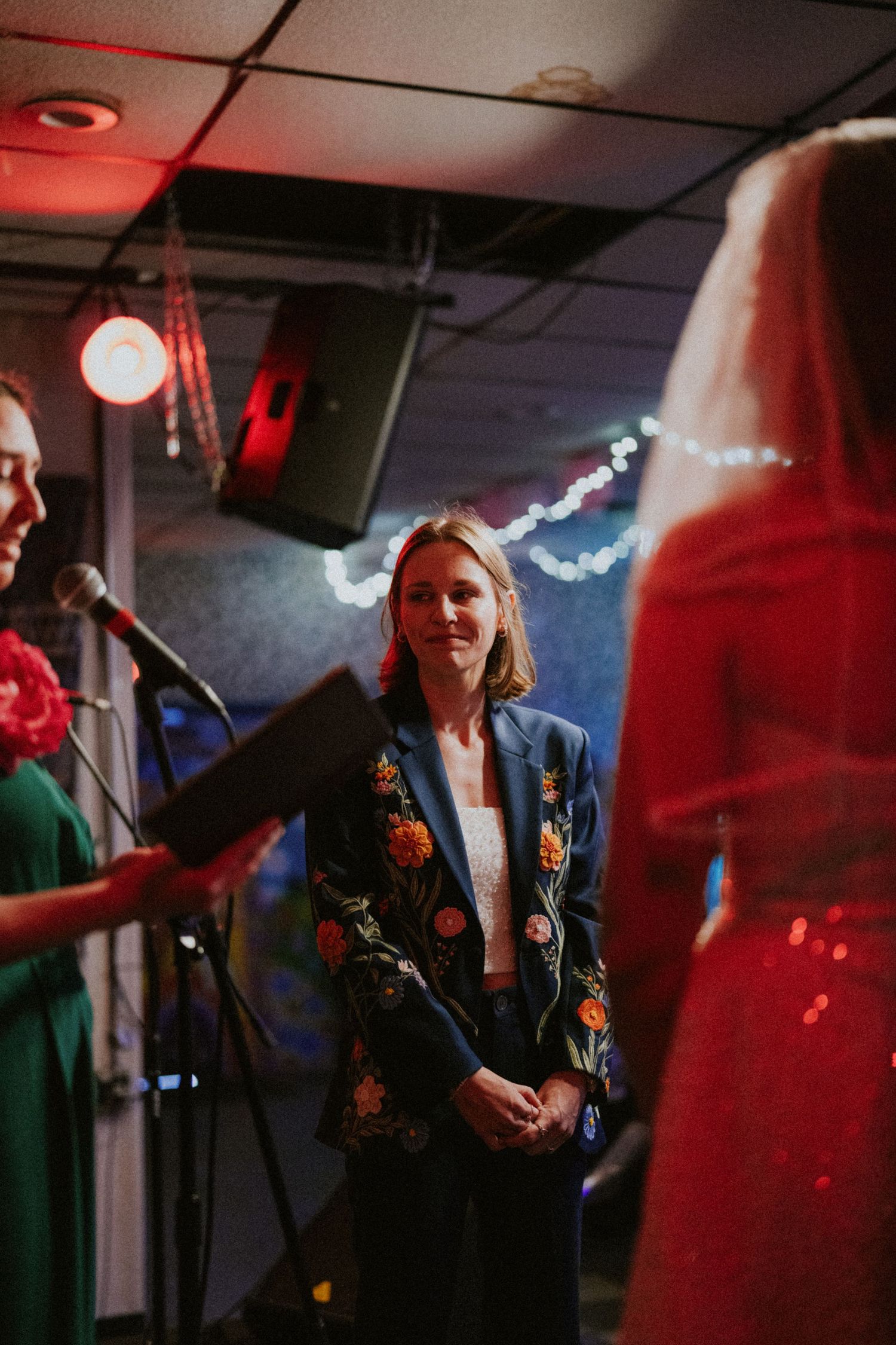 A woman in a floral embroidered blue blazer stands at a wedding ceremony microphone, smiling warmly.