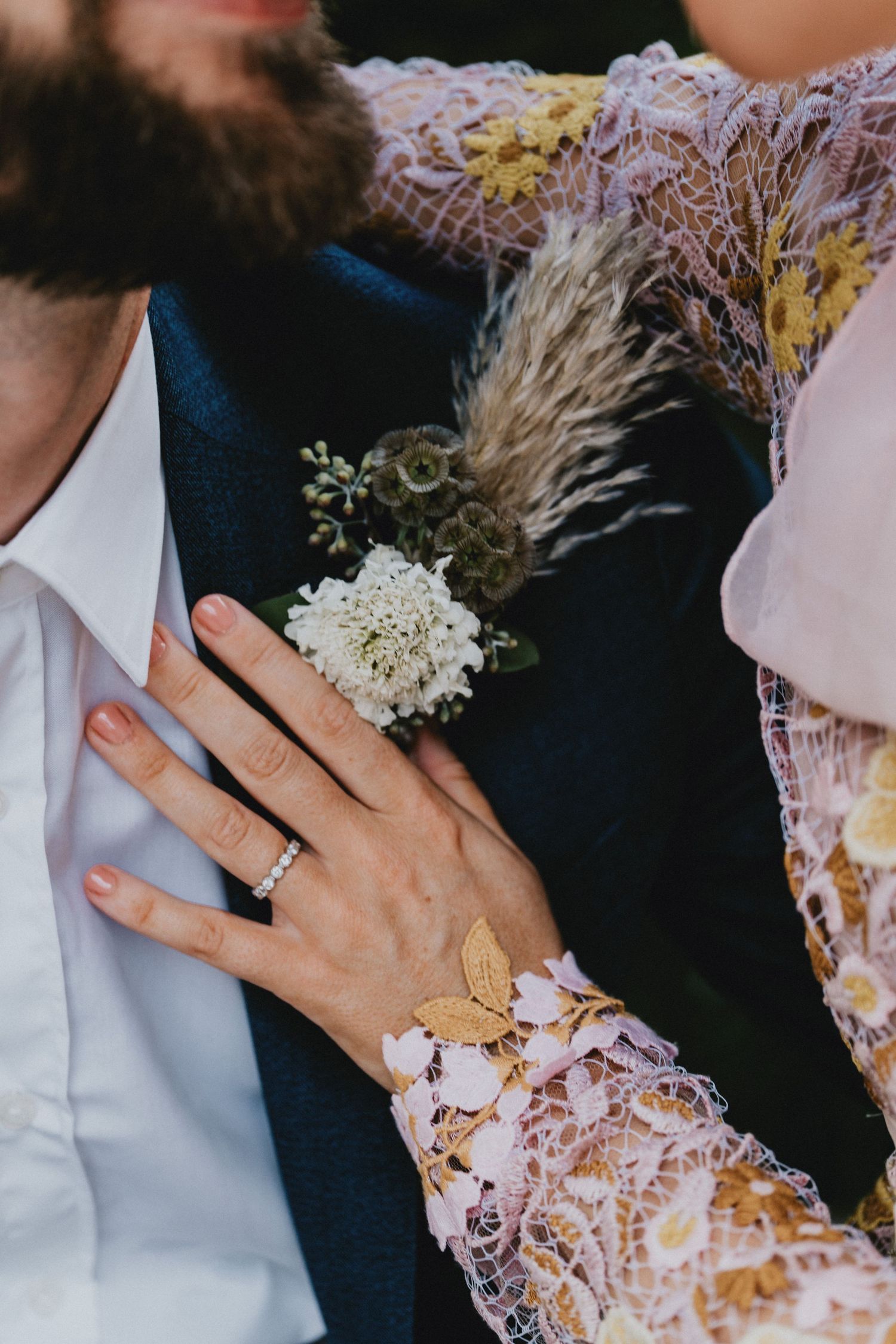 Close-up of bride's hand with diamond ring resting on groom's navy suit with white floral boutonniere.