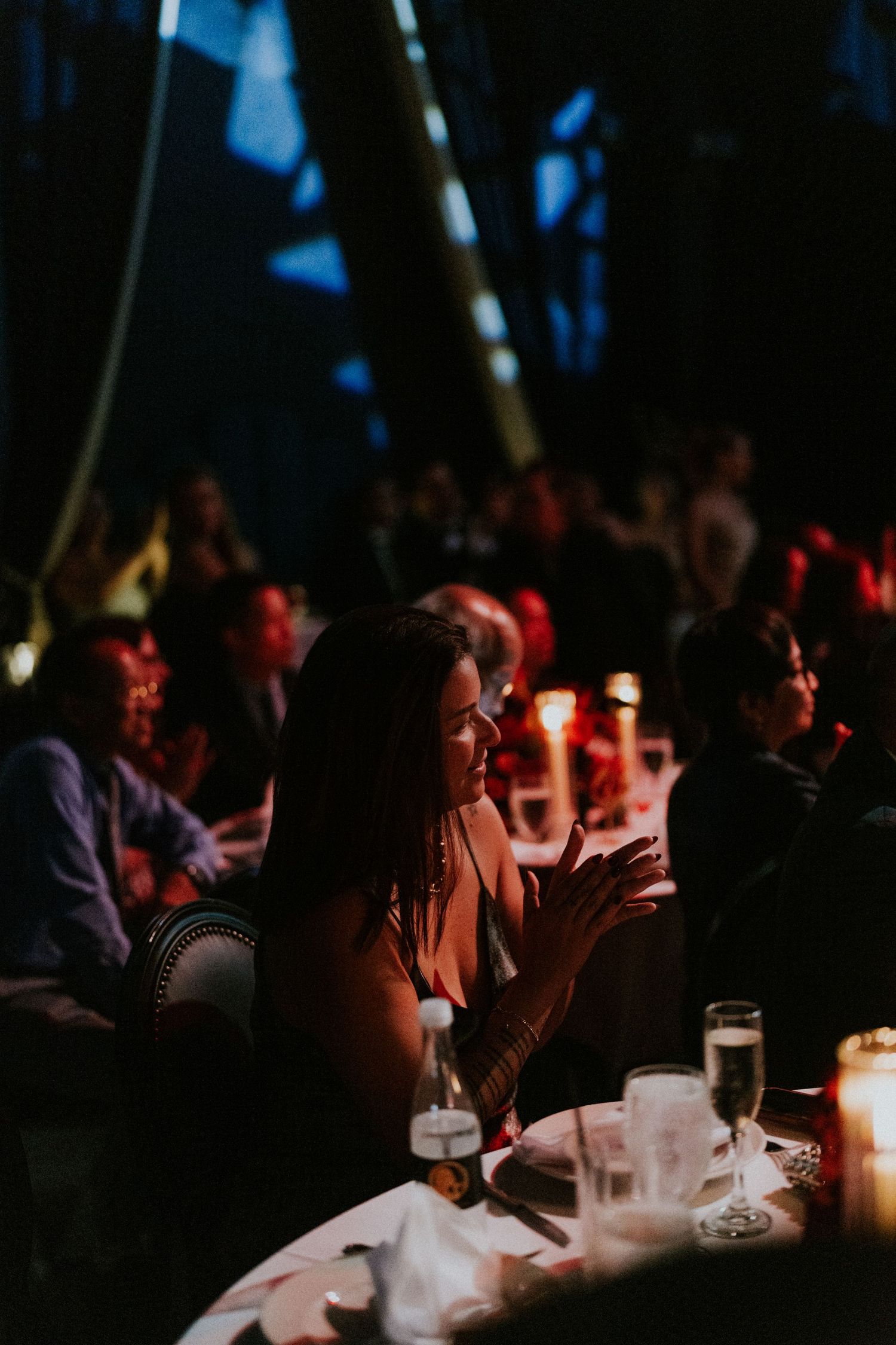 Woman clapping at candlelit gala dinner event with guests seated at tables in dimly lit venue.