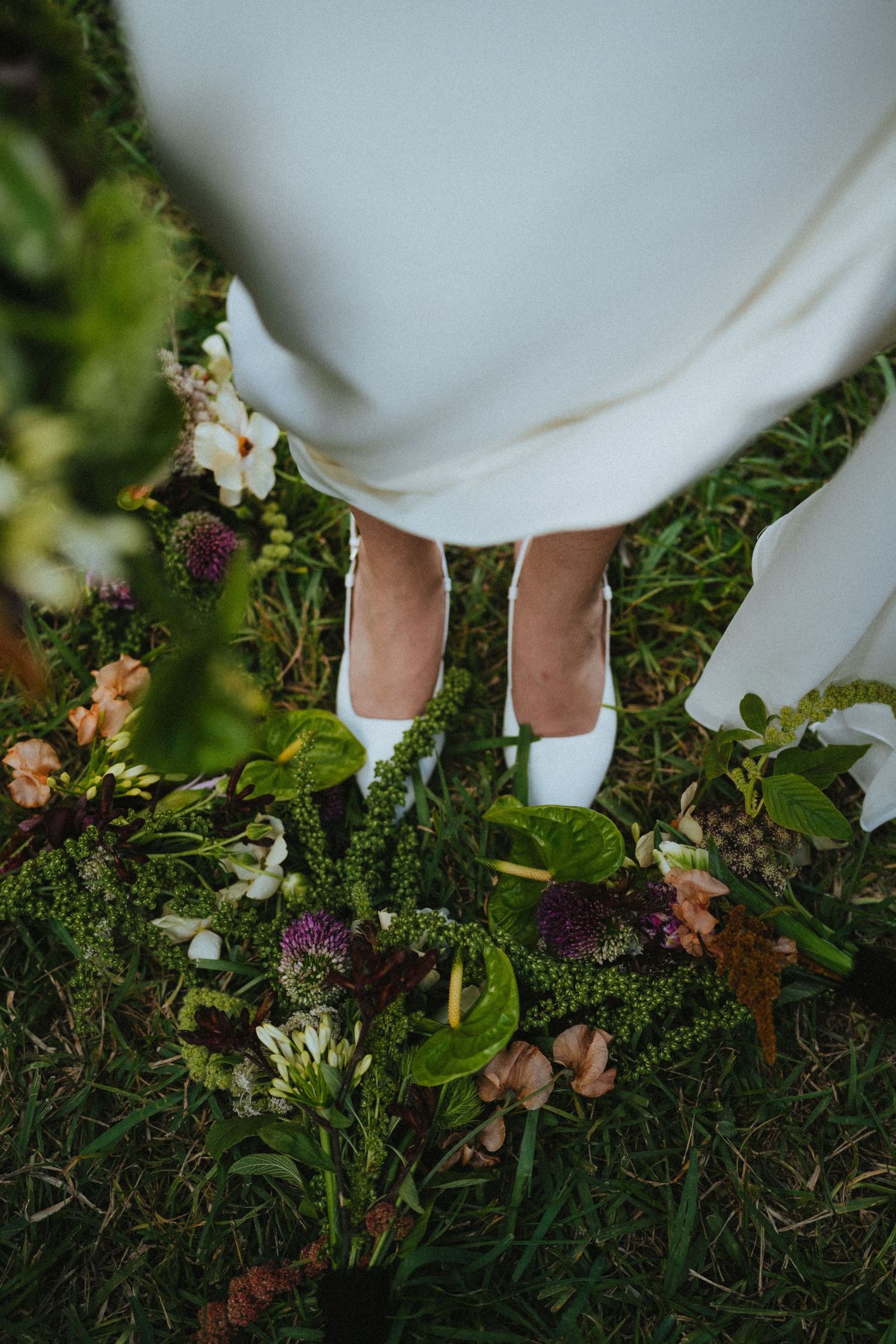 Bride in white dress and white heels standing on grass surrounded by a lush bridal bouquet with purple and green flowers.