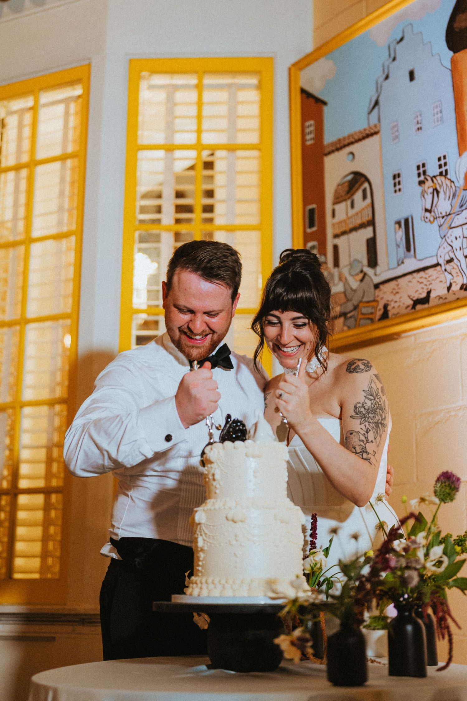 Smiling bride and groom lick frosting from their fingers while cutting a three-tier white wedding cake.