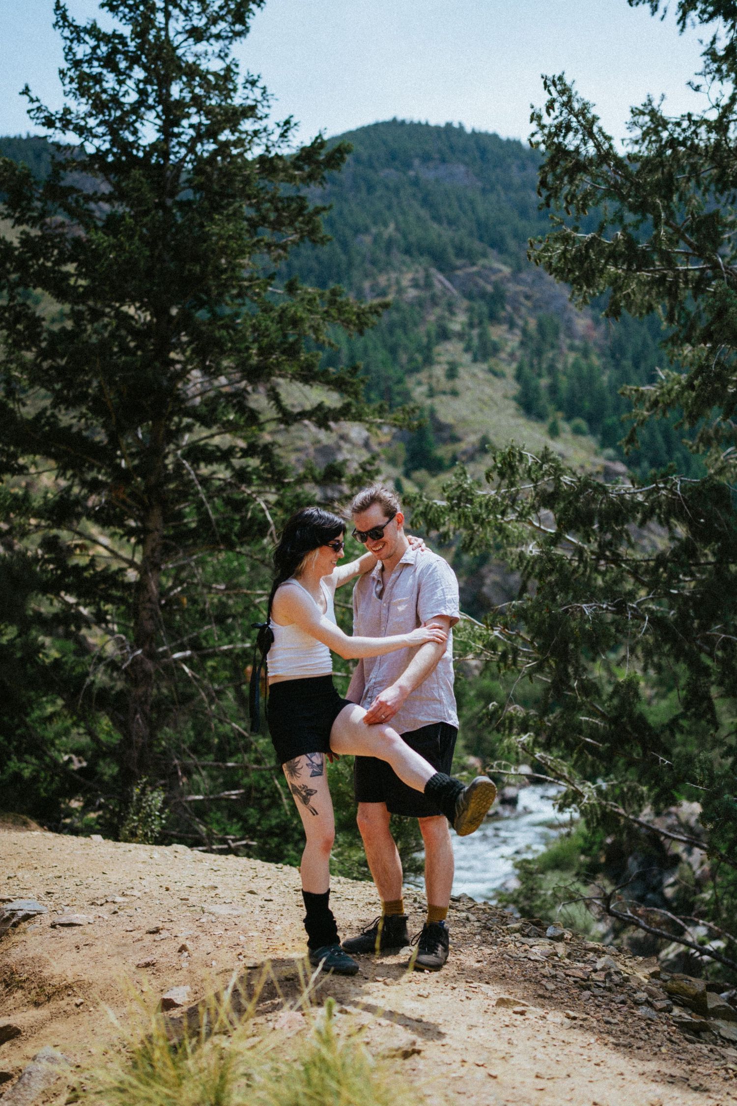 Couple sharing a tender moment on a rocky mountain trail surrounded by pine trees and a scenic river valley backdrop.
