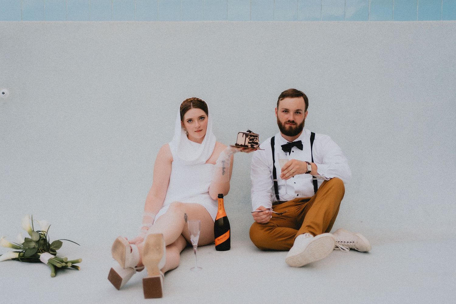 Bride and groom sitting against white wall with champagne bottle and polaroid camera.