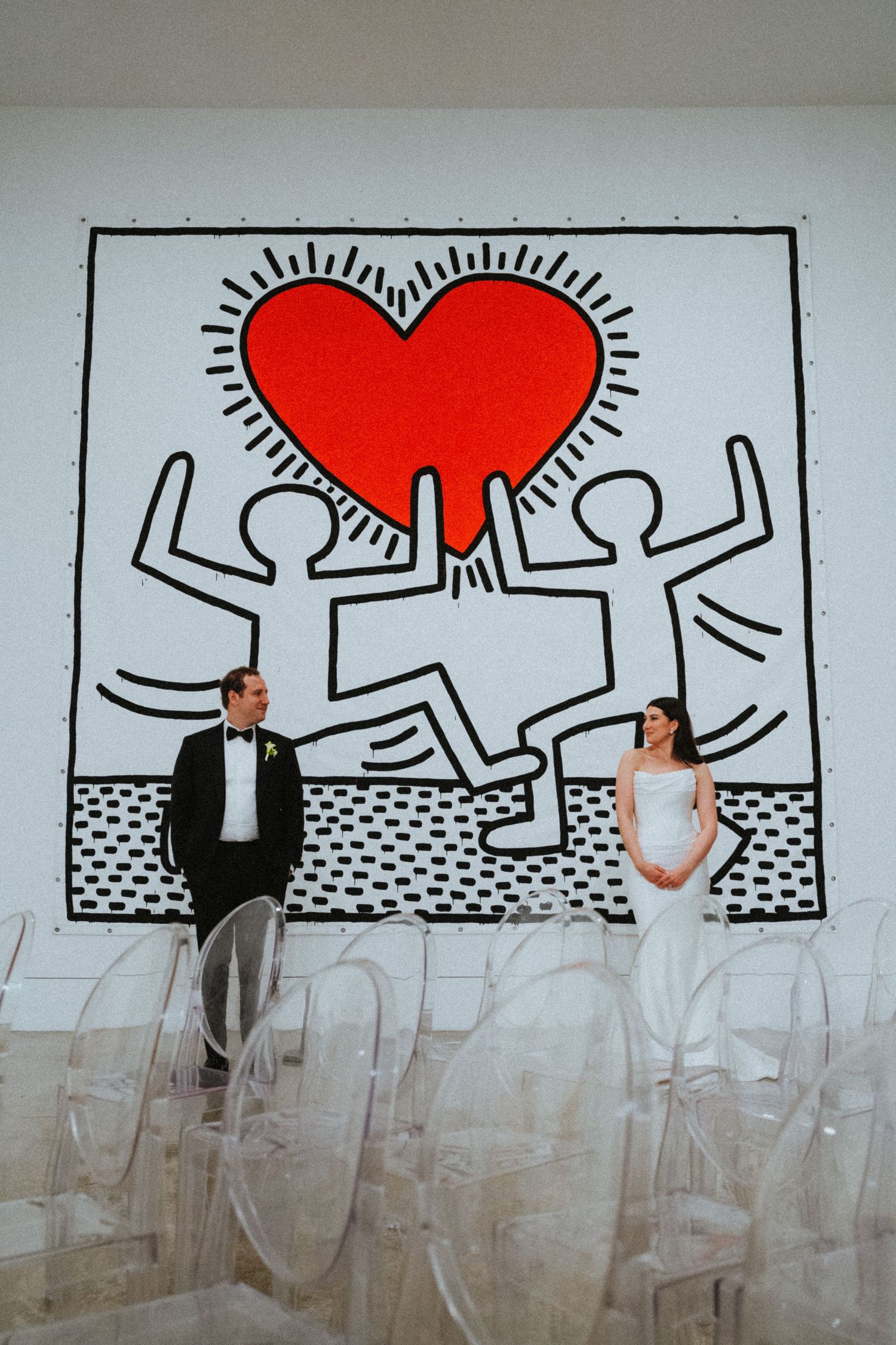Bride and groom posing in front of a Keith Haring heart mural at a modern wedding venue with clear acrylic chairs.