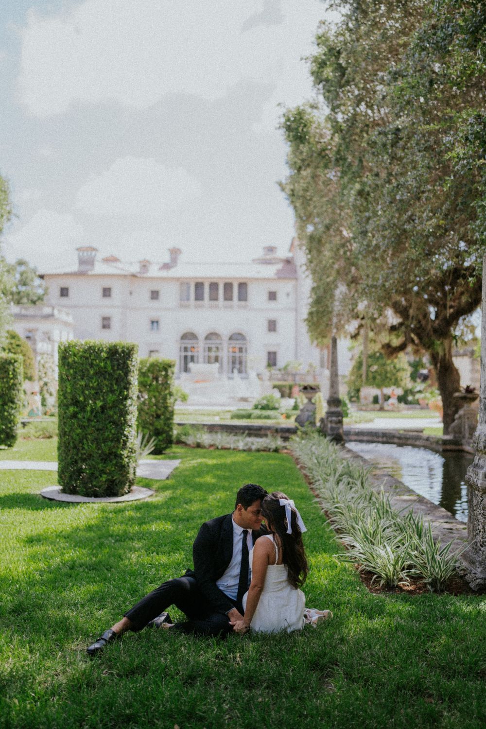 Couple sits on grass in front of elegant white mansion with manicured gardens and water feature.